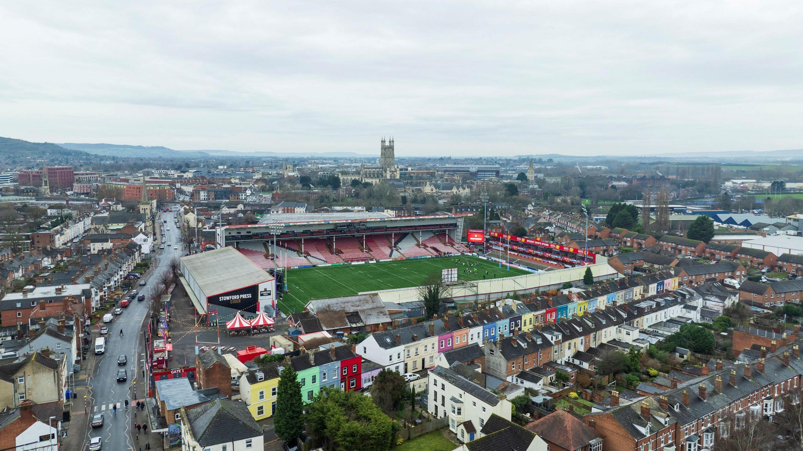 An aerial shot of Gloucester with Kingsholm stadium in the foreground, along with rows of colourful houses, and the city centre in the distance with the spire of Gloucester Cathedral visible