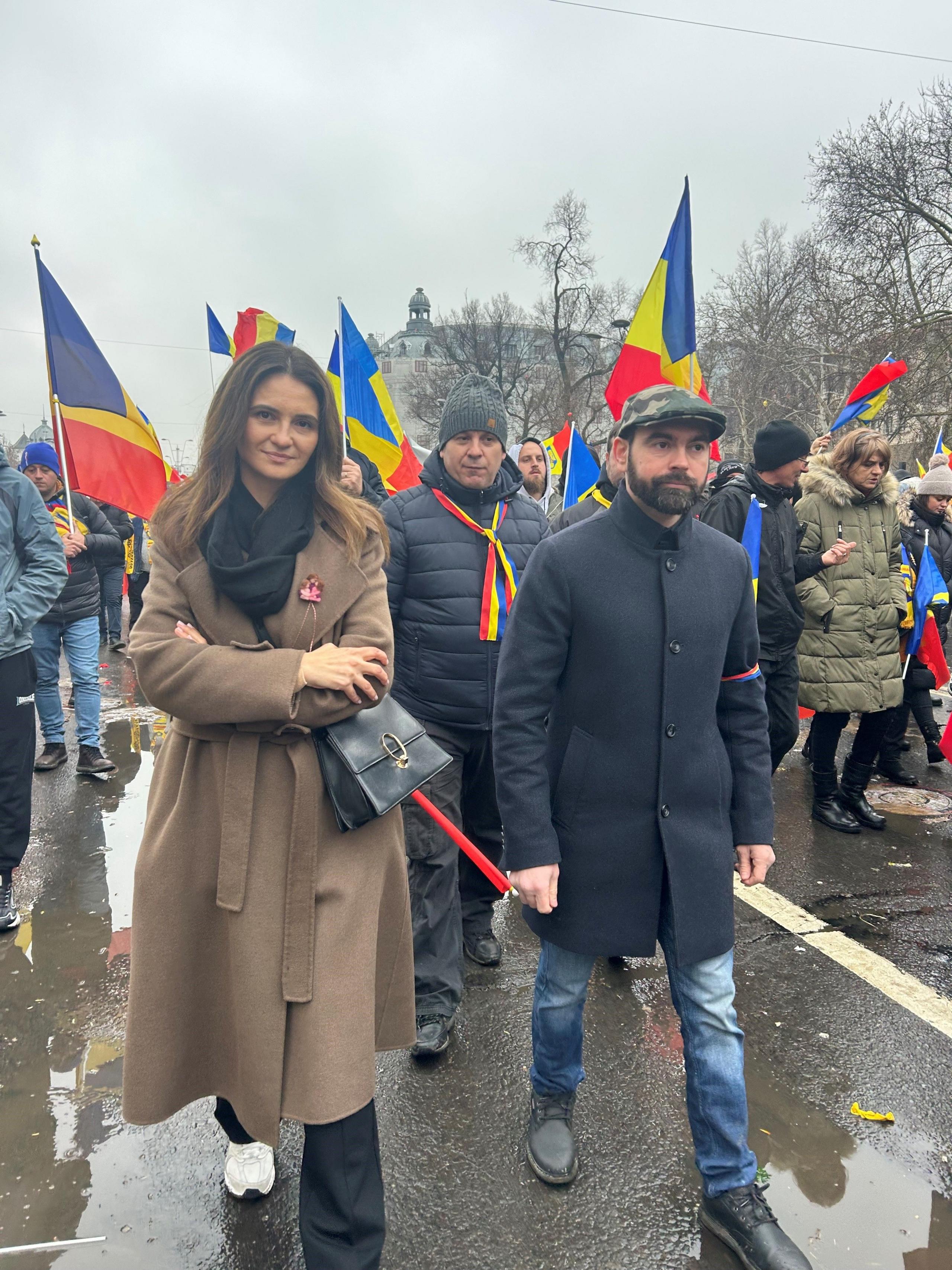 A woman in a brown trench coat marches in a large group, many of whom are carrying the Romanian flag. She is smiling to the camera.