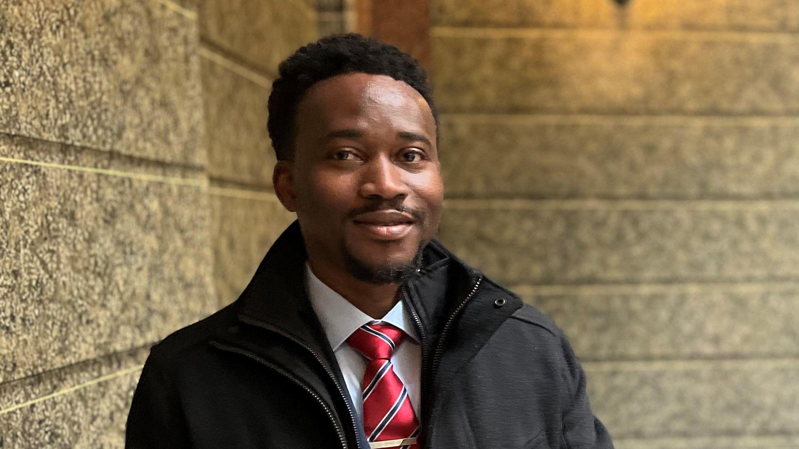 Mohamed Wurie smiles as he stands with his hands clasped together, with a staircase at St George's Hall behind him. He is wearing a dark blue suit with a white shirt and red, white and blue striped tie, along with a black coat.