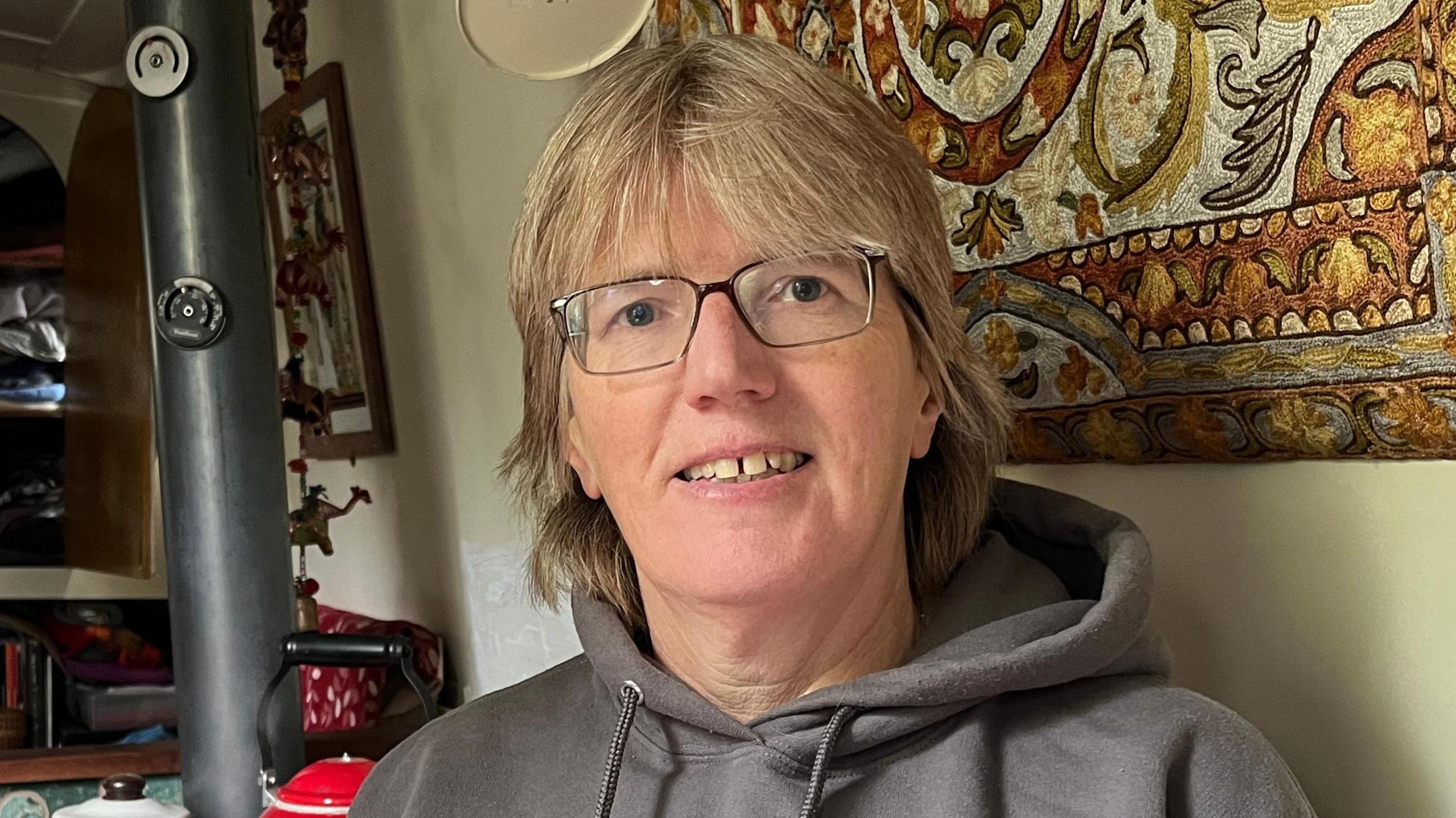 Ally Clarke is sitting inside her canal boat which has cream walls. Behind her is a tapestry, a stove and a kettle. She has medium length blonde hair and is wearing glasses and a grey jumper with 'this is how i roll' printed on the front.