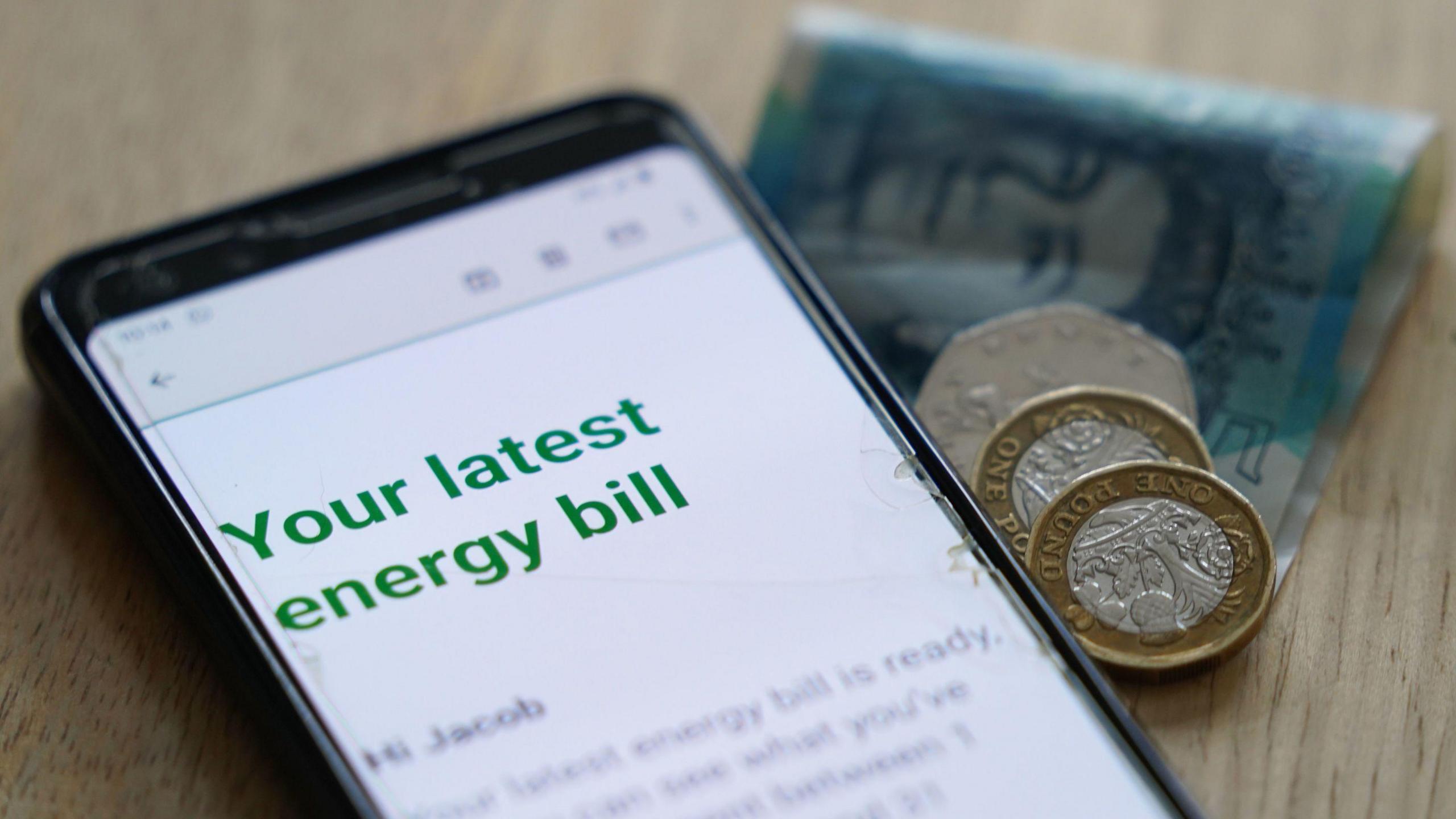 A phone screen reading 'Your latest energy bill'. A five pound note, two pound coins, and a 50p coin are next to it.