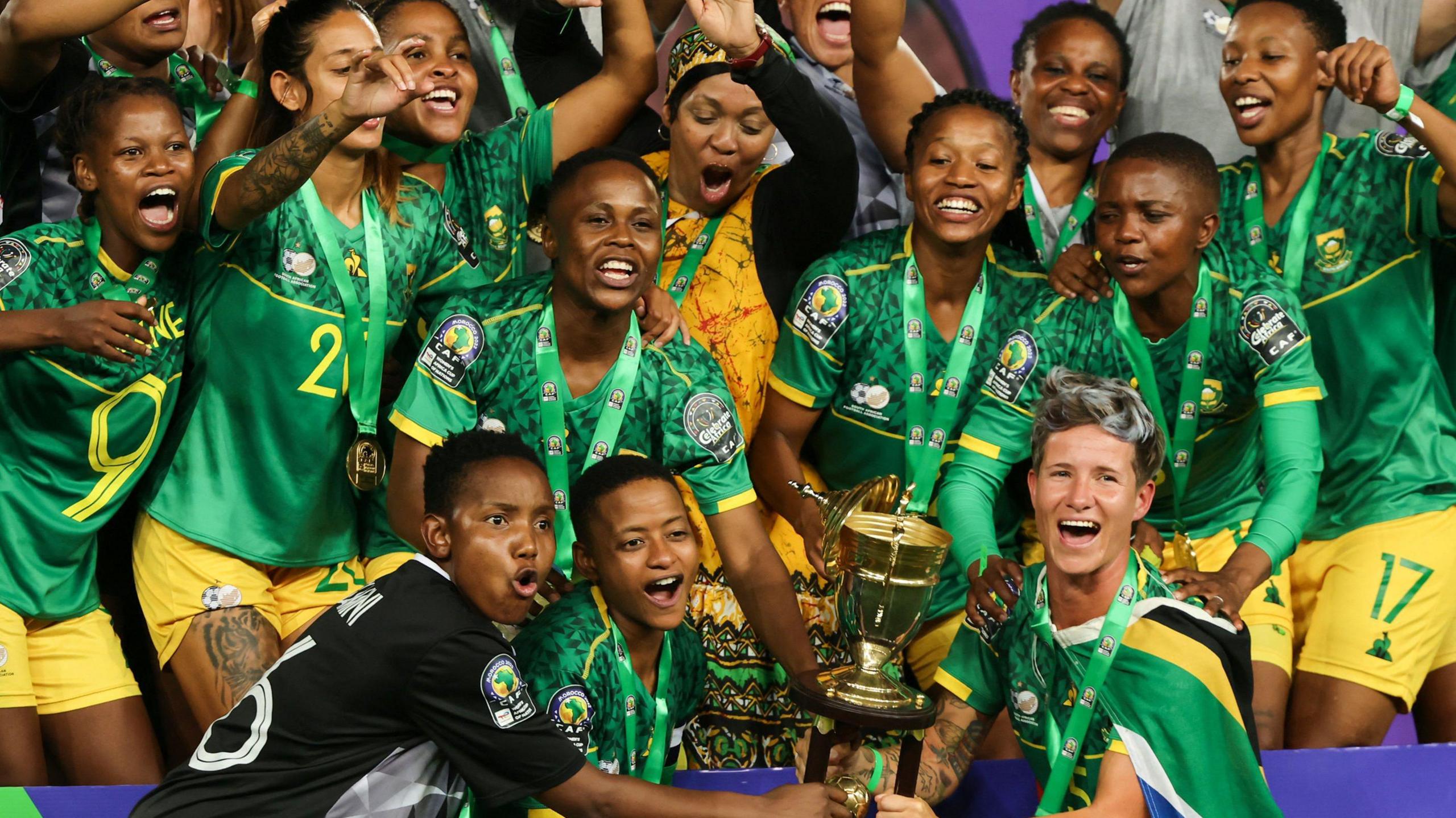 A jubilant South Africa team, some with medals around their necks on green ribbons, pose with the Women's Africa Cup of Nations trophy in 2022