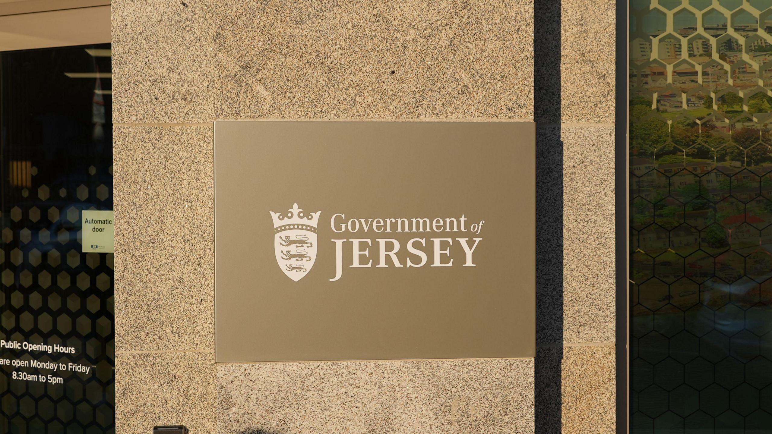 A golden Government of Jersey sign with the Jersey emblem to the left of the wording put on a granite wall with an automatic door on the left and glass window on the right.