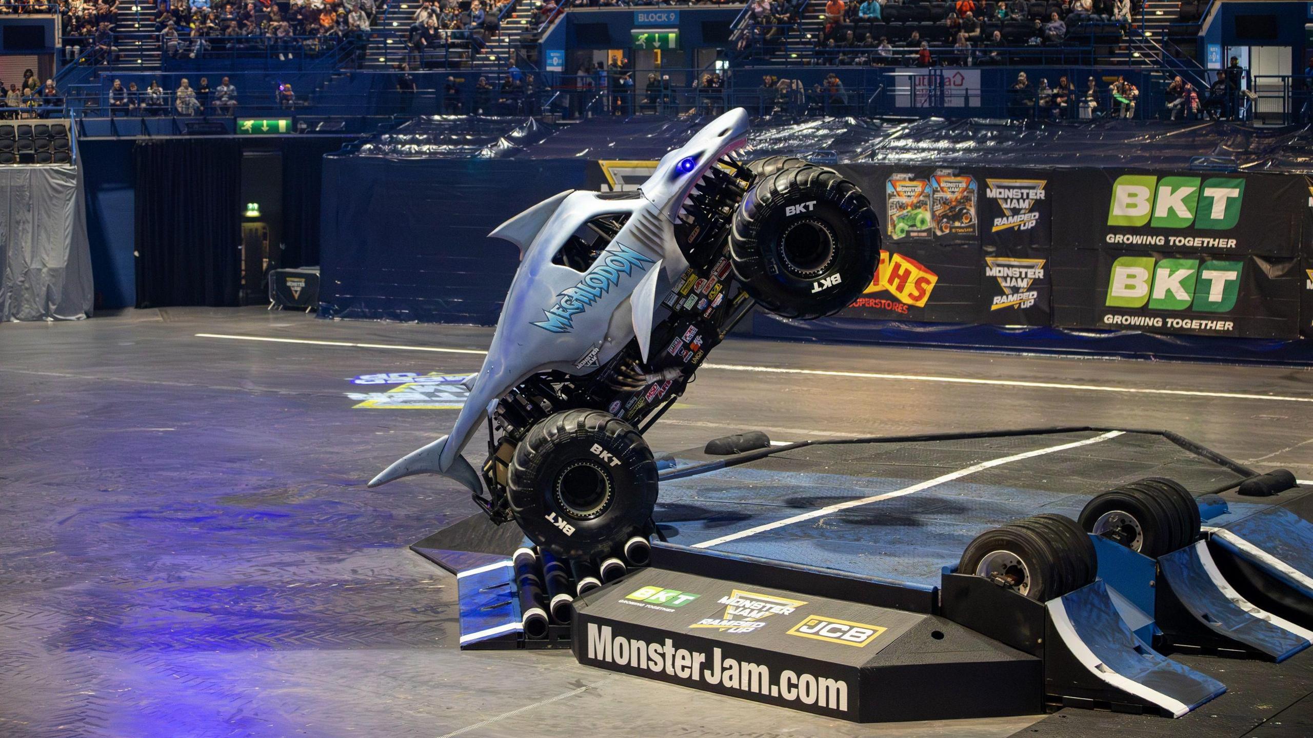A monster truck doing a wheelie over a large ramp in an arena. Fans are in the stands. The truck is decorated to be the shape of a large shark, with a glowing blue eye and sharp teeth. On its side in blue writing, it reads "megalodon"