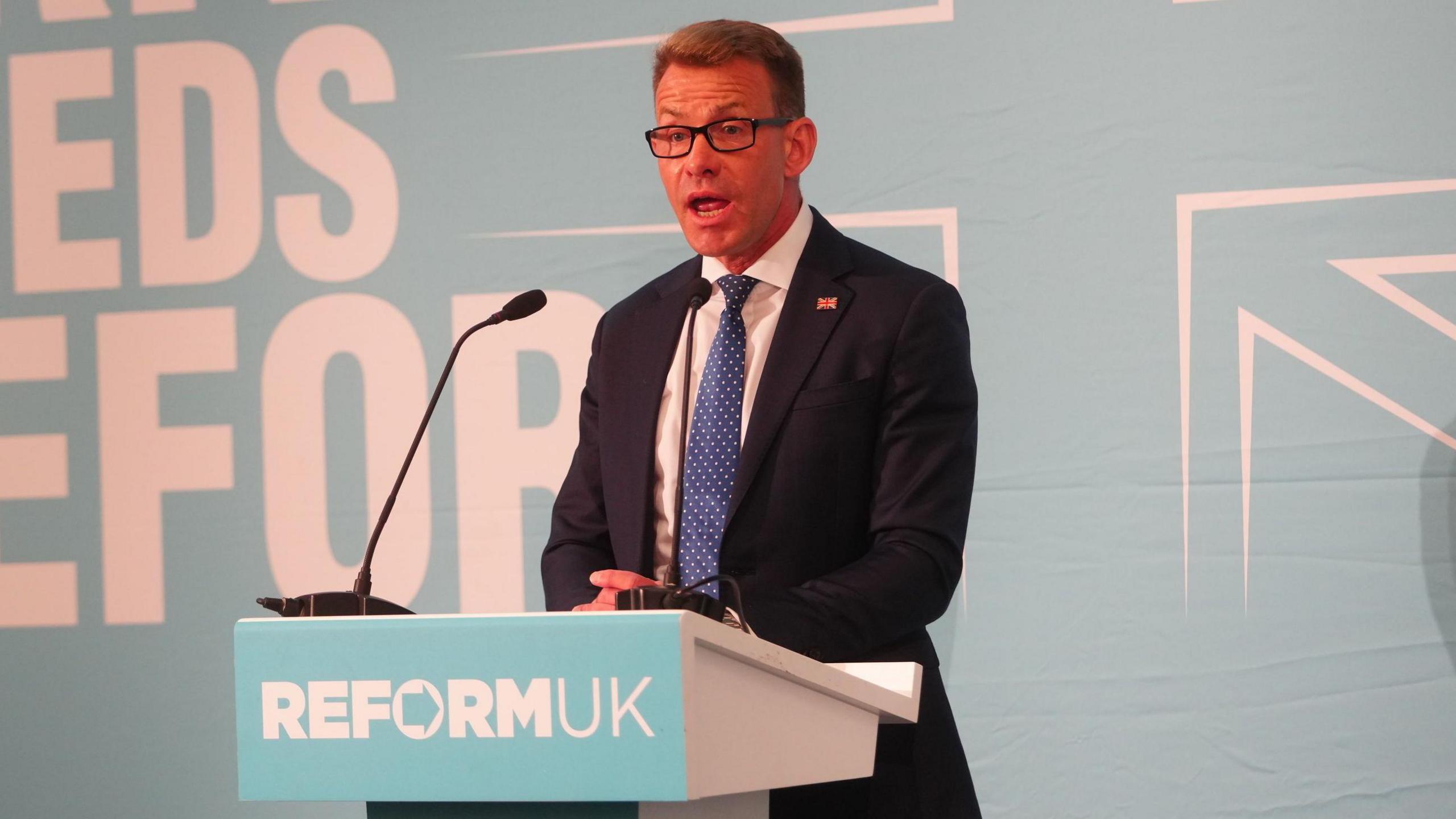 David Bull gives a speech behind a blue podium, reading "Reform UK".