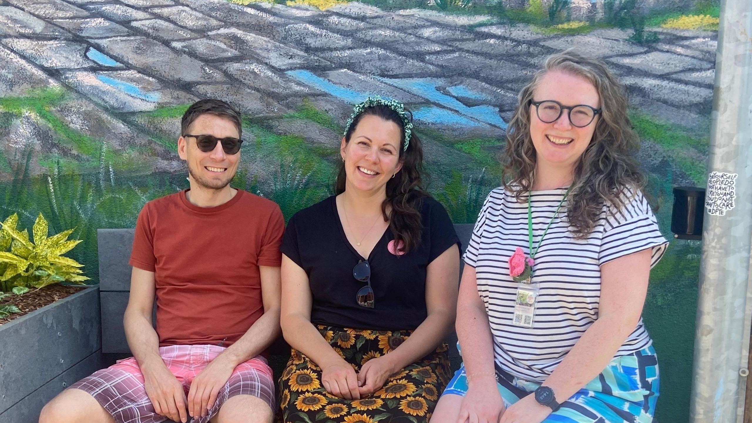 Three people, two women and a man, sit on a wooden bench under a mural of cobbles and grass. They are all smiling at the camera and wearing summery clothes.