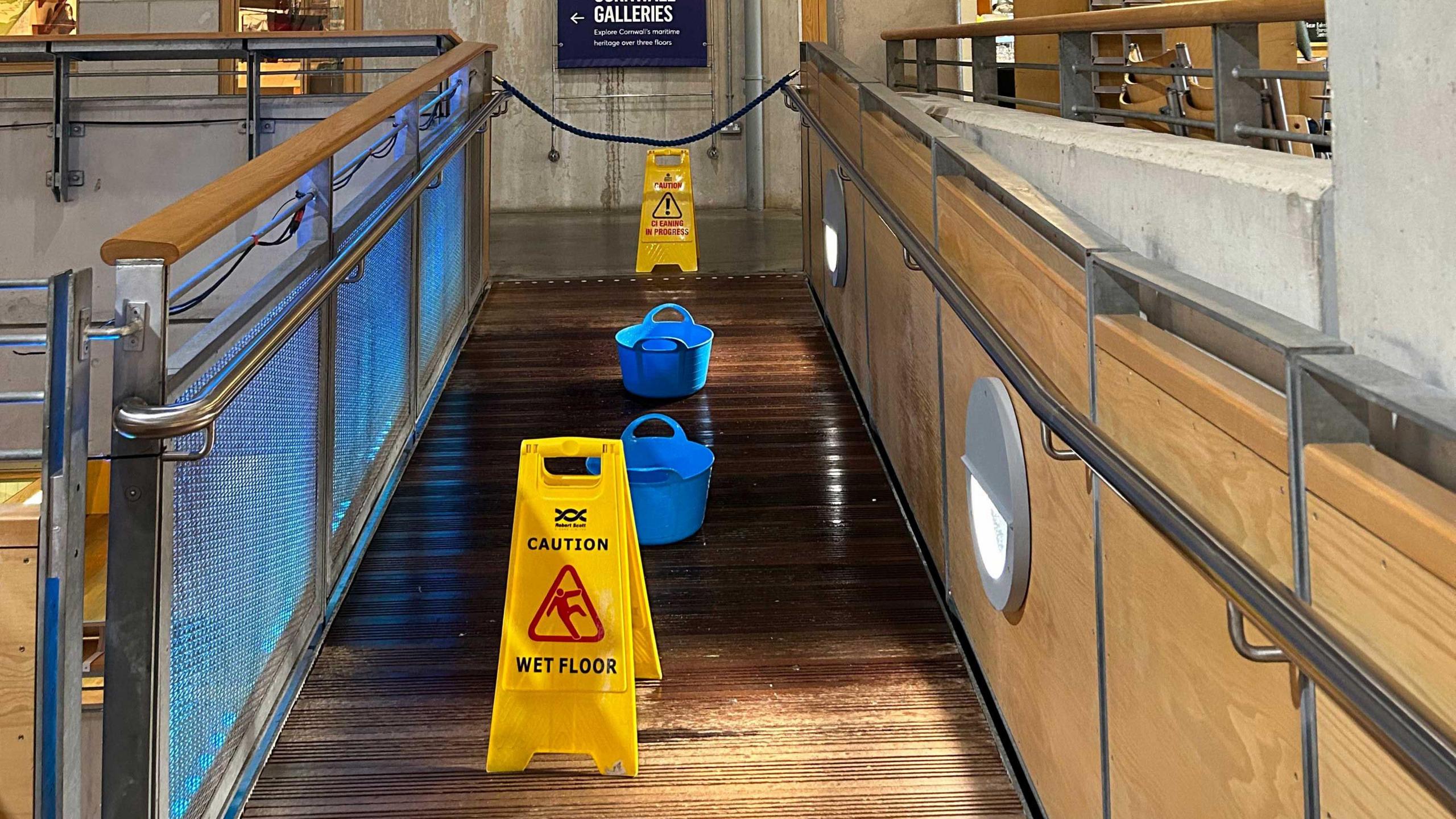 A pathway at the museum is photographed with yellow caution signs indicating a wet floor. There are also blue buckets scattered across the path collecting water.