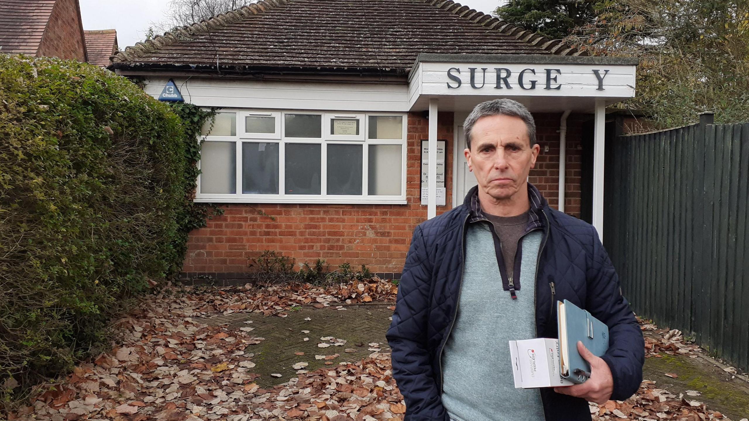 A man looking pensive holding books and papers under his arm, in front of a run down looking GP practice with the sign above the door missing the second R from the word SURGERY