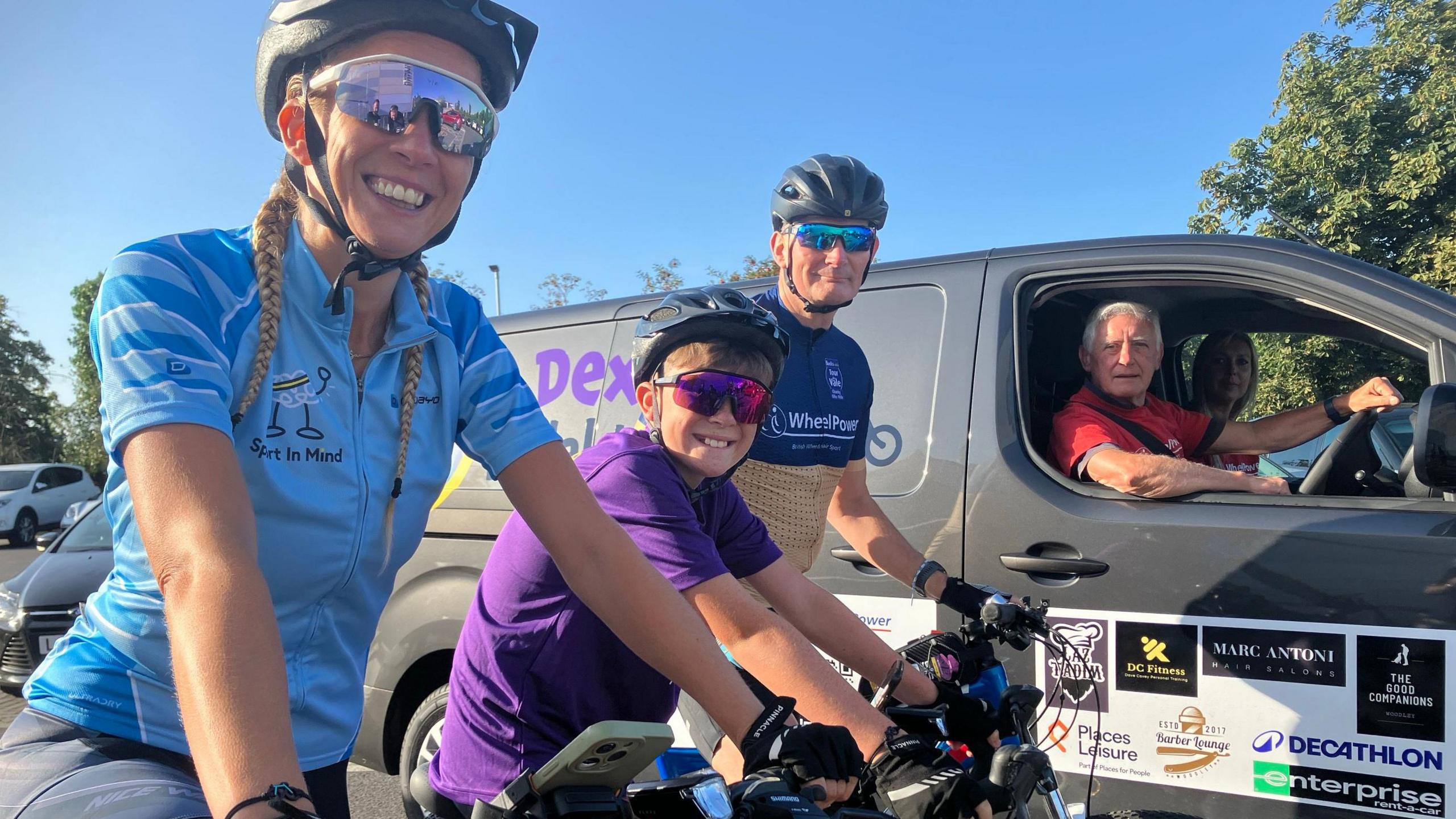 From left to right - Dexter's mother Sam, Dexter and coach Dave Covey as smiling as they are prepared to set off. All three are on their bikes. Sam wears a blue top for Sport in Mind, Dexter wears a purple T-shirt for Falconi Hope, and Mr Covey wears a dark blue and yellow top for WheelPower. A driver in a van with Dexter's name on it is next to them. A woman can be seen in the passenger seat. It's a sunny day.