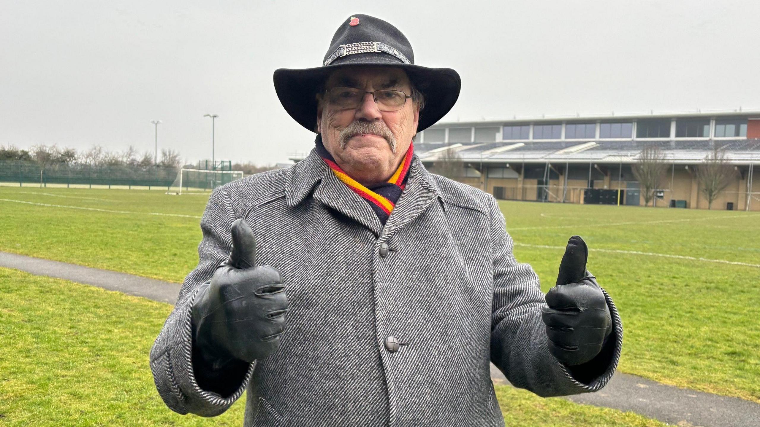 A man with a moustache is wearing a black hat and grey coat. He stands in front of a grass football pitch and has his thumbs up.