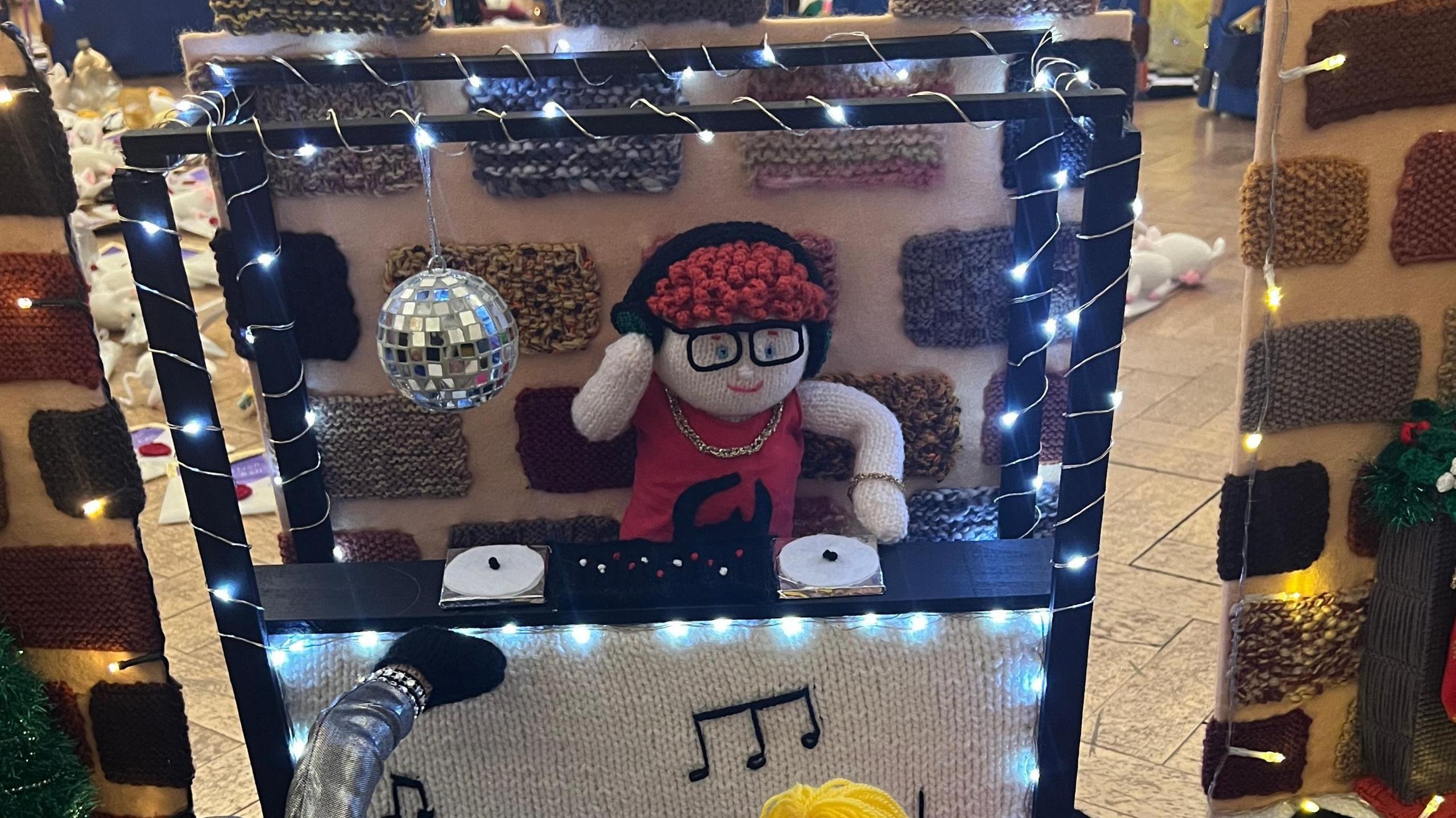 A knitted DJ in a booth, complete with headphones, turntable, lights and glitter ball. The DJ has red curly hair and has glasses on his face. He has a necklace and red top on. 