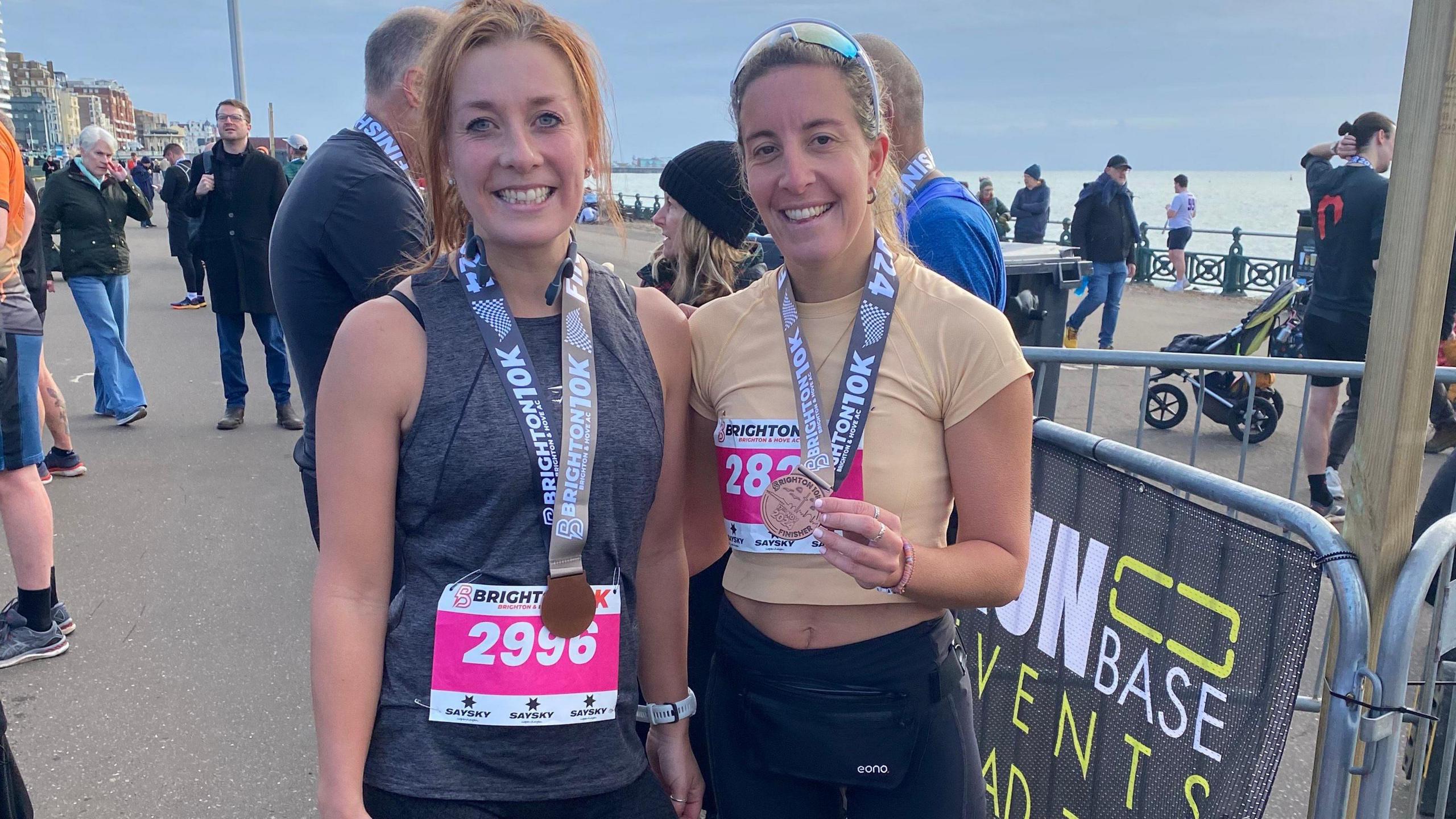 Two women standing by the seafront holding their running medals. 