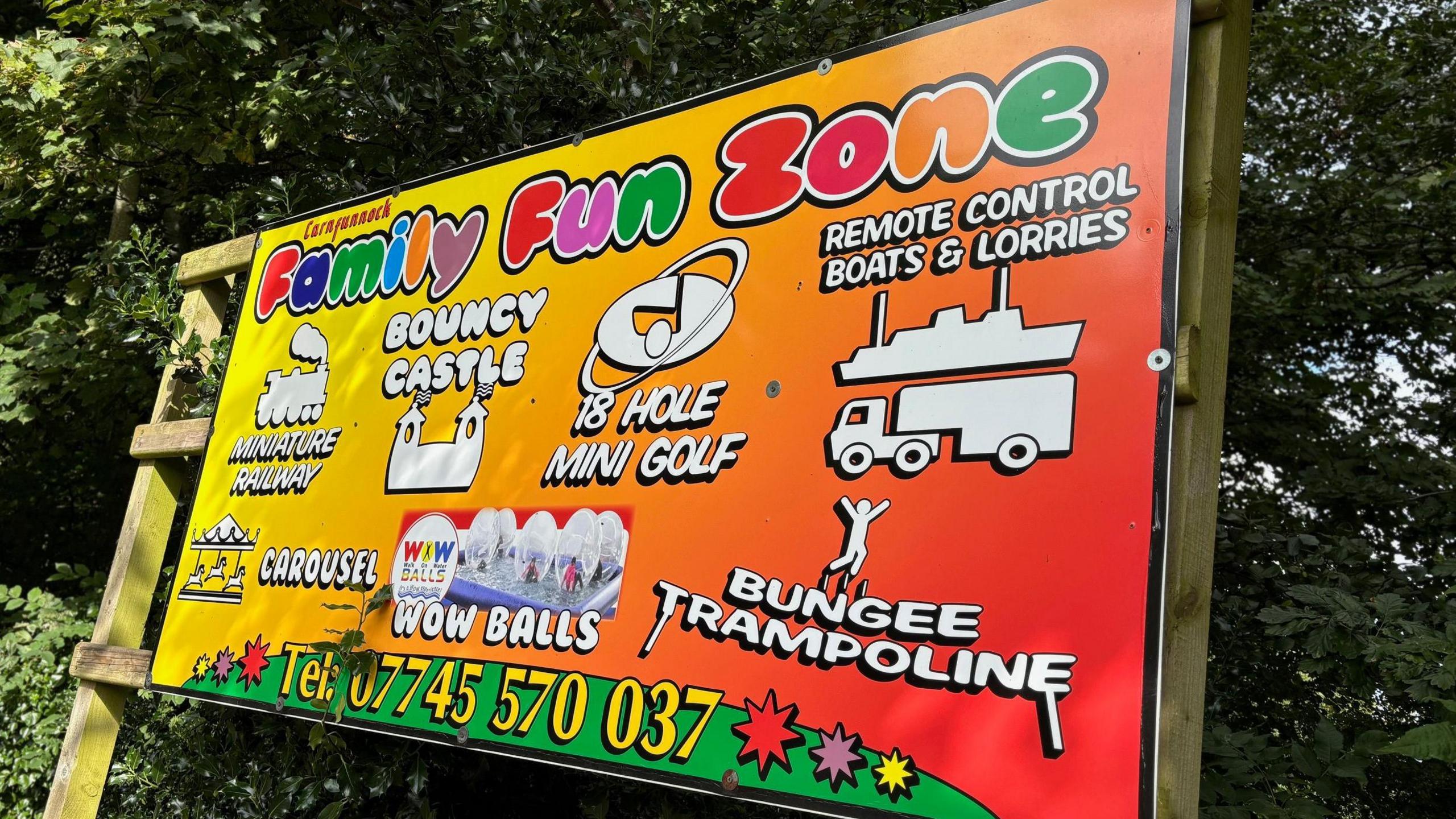 Business sign for the family fun zone at Carnfunnock Country Park - it is red and yellow with Family Fun Zone emblazoned at the top in numerous colours. It advertises Bungee Trampoline, Wow Balls, Bouncy Castle, 18-hole mini golf, carousel, miniature railway, remote control boats and lorries. 