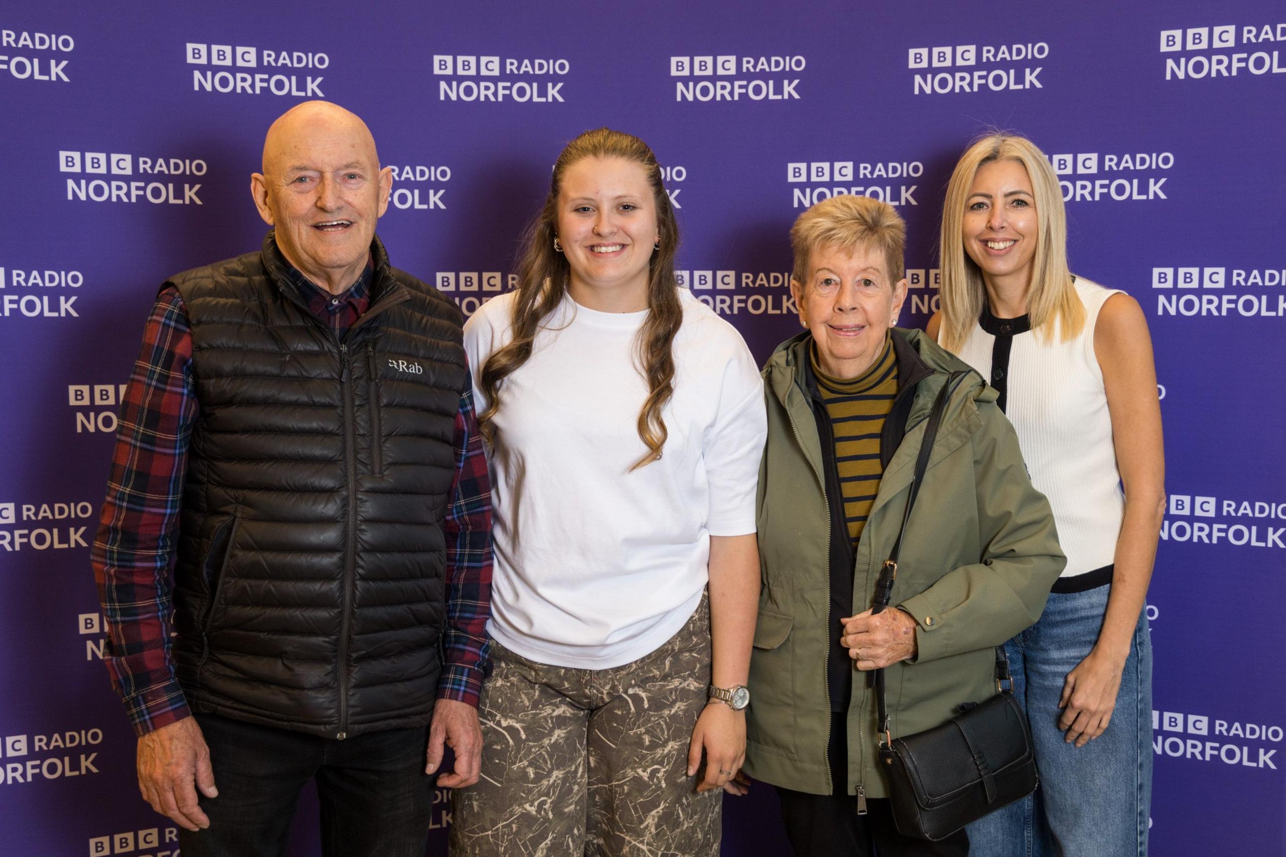 Three women of varying ages and one older man stand smiling in front of purple BBC Radio Norfolk branding.