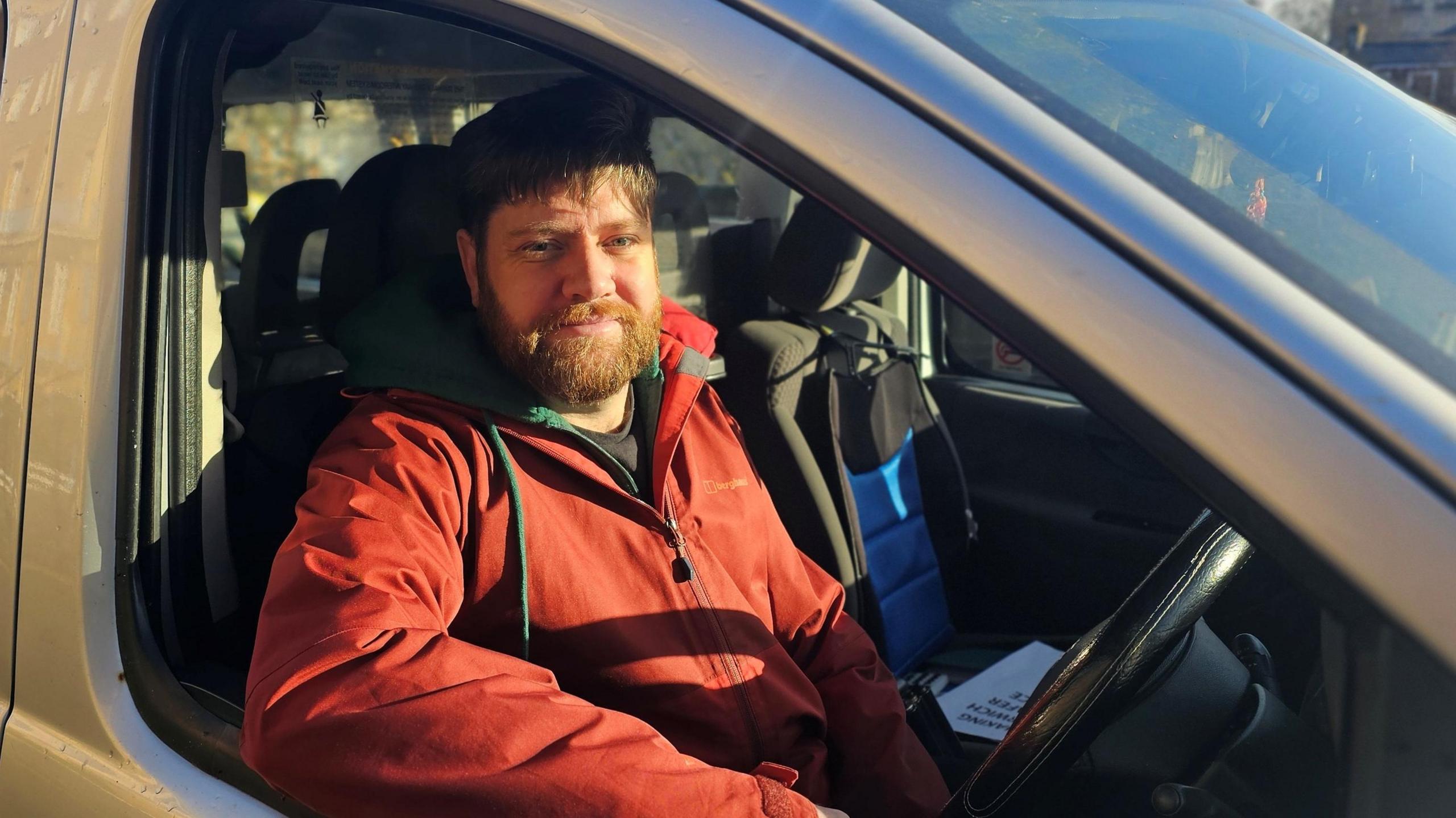 Kierran Greaves sitting in the driver's seat of a hackney carriage. He is wearing a red coat over a dark top