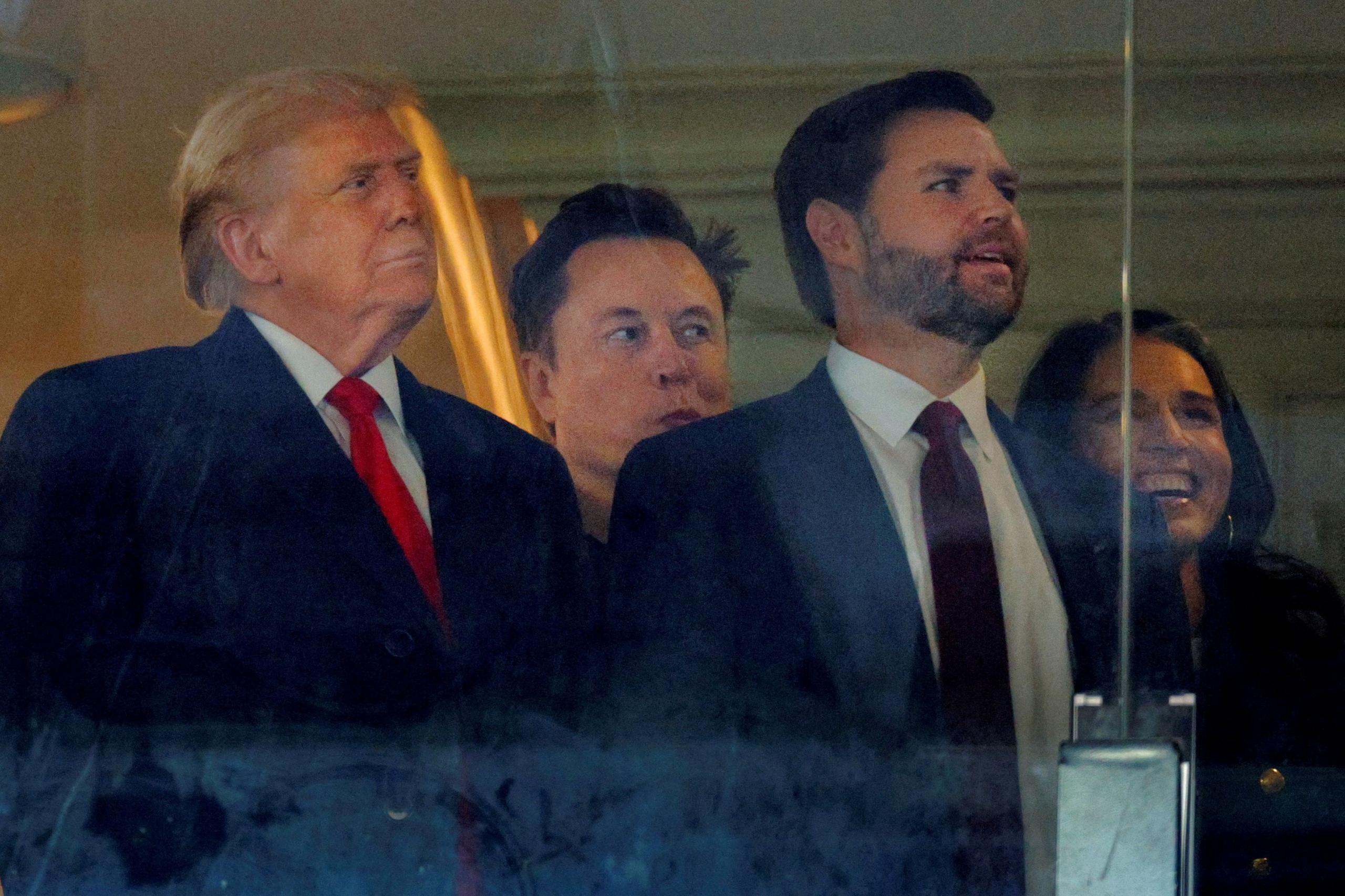 Trump wears and dark coat and a red tie, he is stood next to Vance who is also wearing a dark-coloured suit. Between their shoulders can be seen the head of Elon Musk. All three are looking to their left