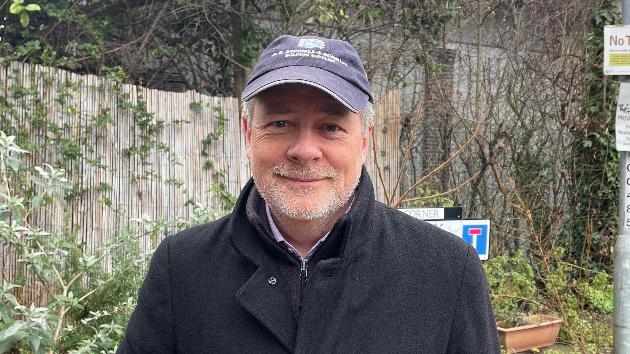 Richard Darler wearing a cap and a coat, smiling at the camera. In the background is a fence separating the street from the Beehive Centre, with overgrown foliage on it.