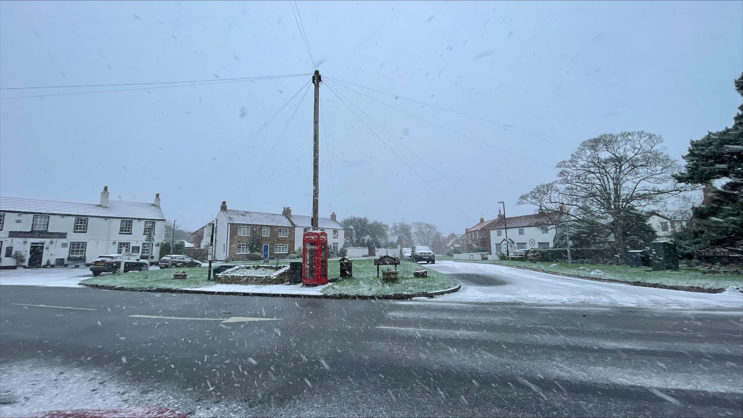 Snow falling on a landscaped village green with a red post box on it. 