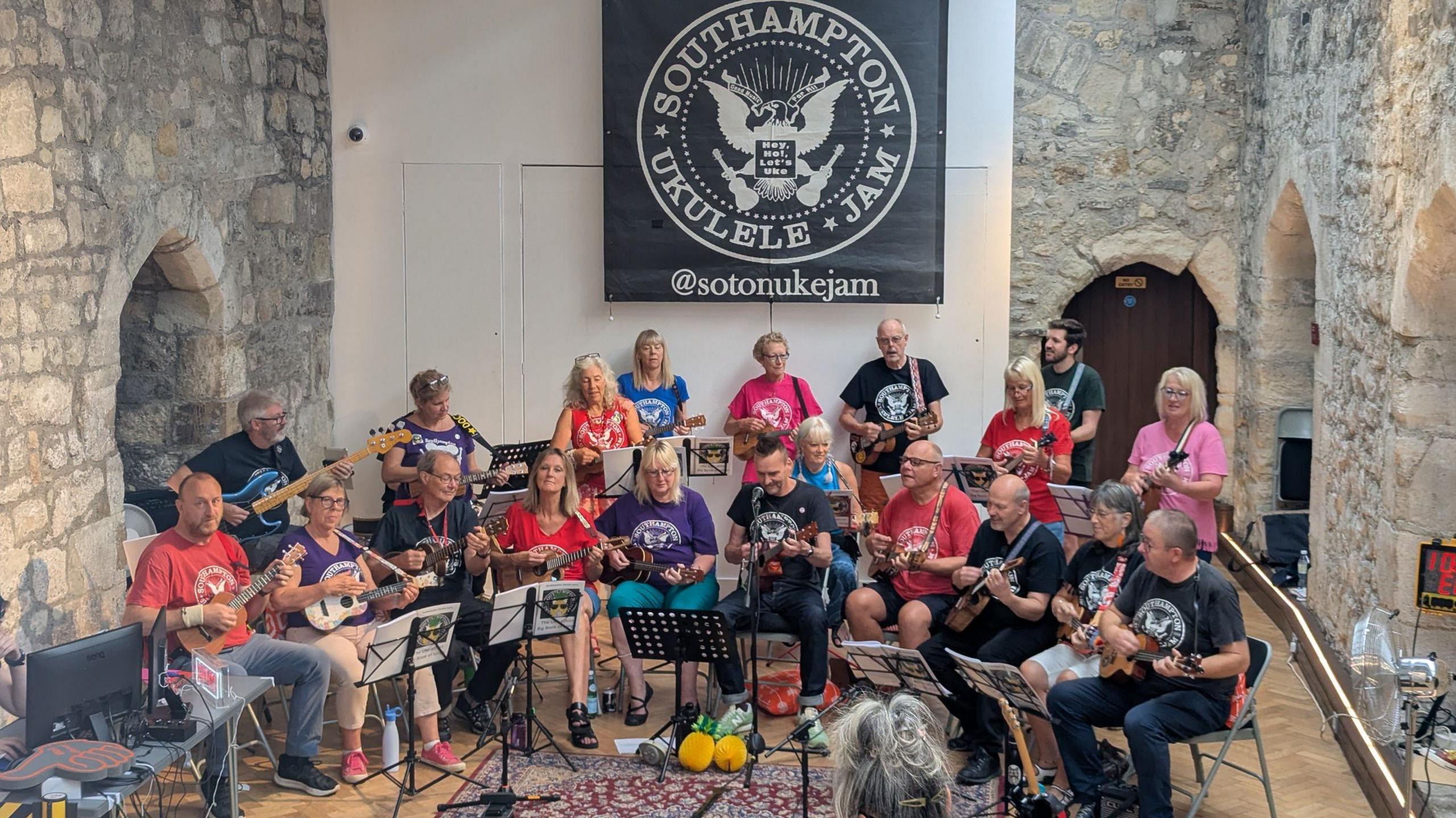 Group of musicians sat in a curve inside a stone building all playing a ukulele .