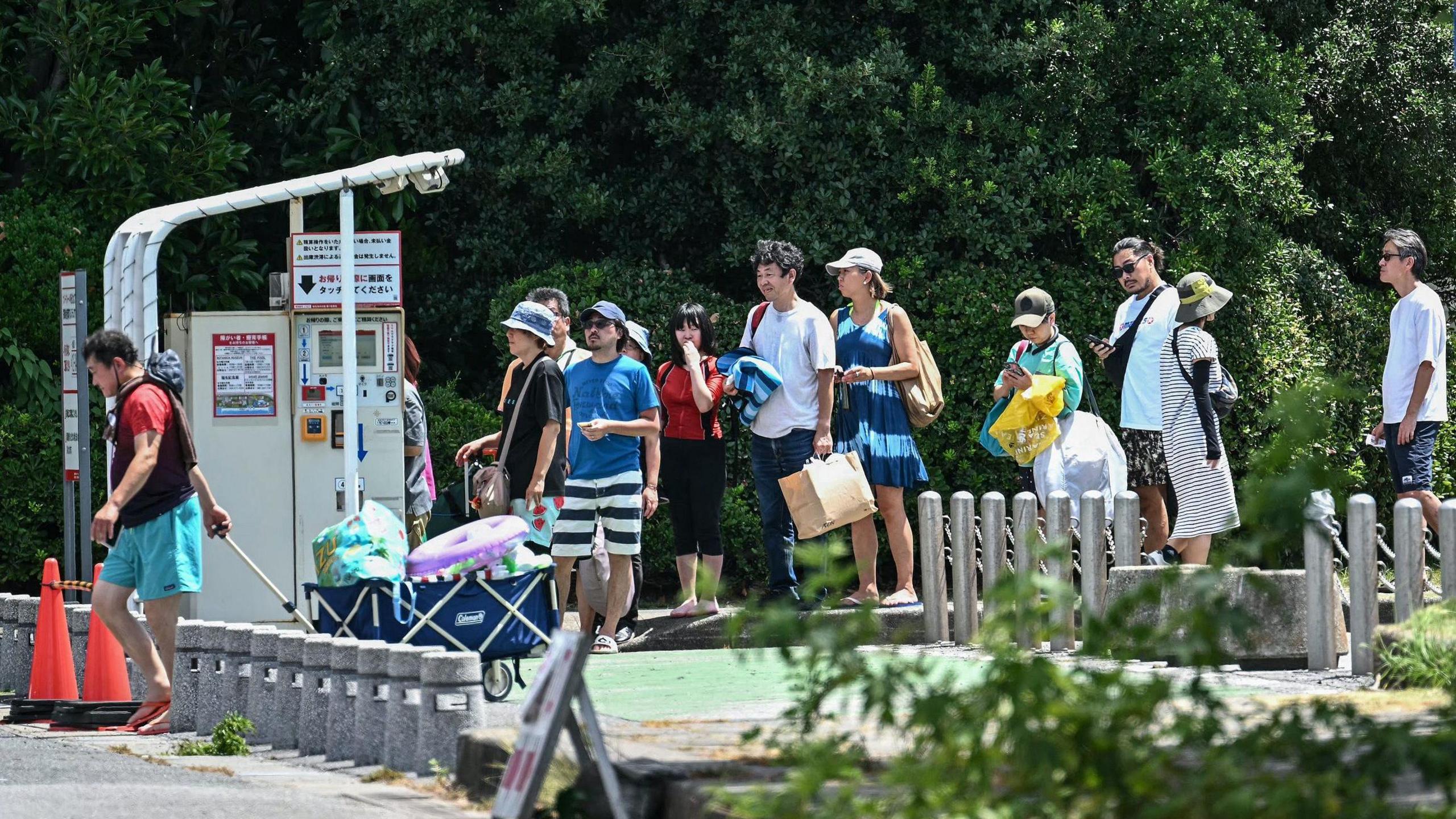 Holidaymakers in beachwear are seen queuing for a ticket machine to pay for parking as they begin to evacuate a holiday resort in Tokyo Bay. There are trees in the background