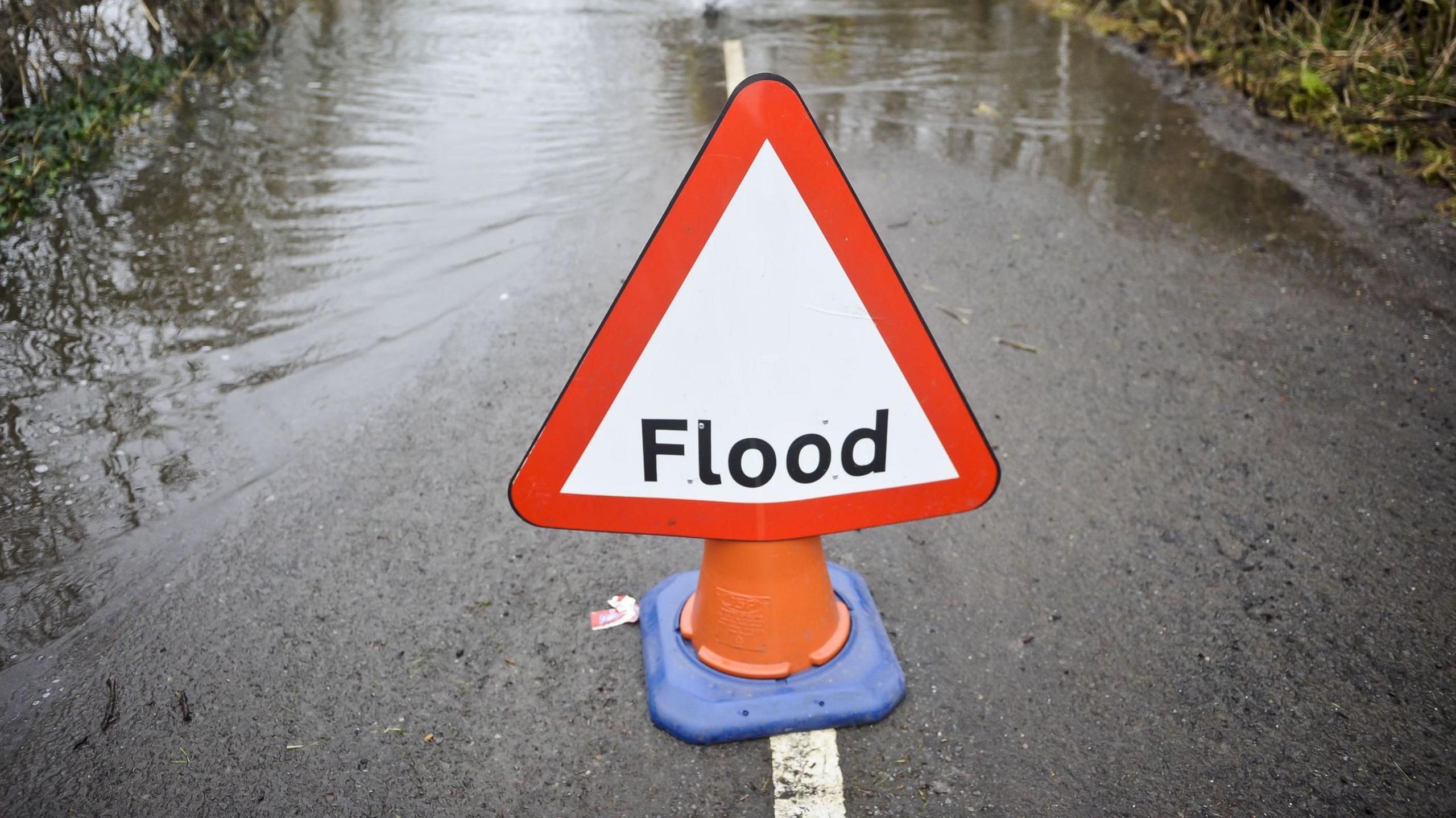Flood warning sign on traffic cone in front of flooded country road 