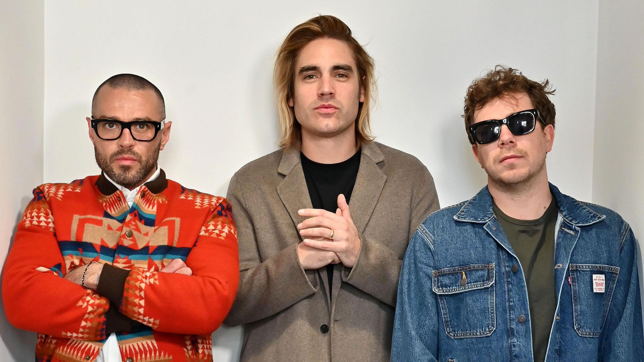 The pop-punk band Busted looking straight at the camera. Matt Willis is wearing black glasses and a red patterned jacket with his arms folded. Charlie Simpson is wearing a black top and beige coat. James Bourne is wearing a green top, denim jacket, and black sunglasses.