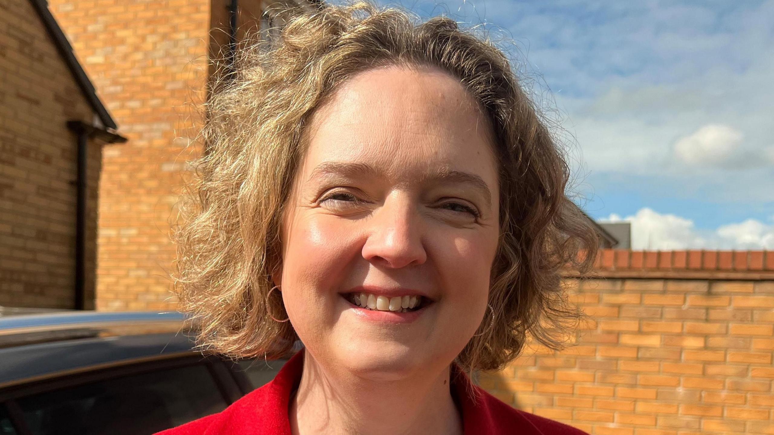 Anna Sabine is smiling at the camera. She is standing in front of a brick wall and there is a parked car to the left of her. She is wearing looped earrings and a red coat.