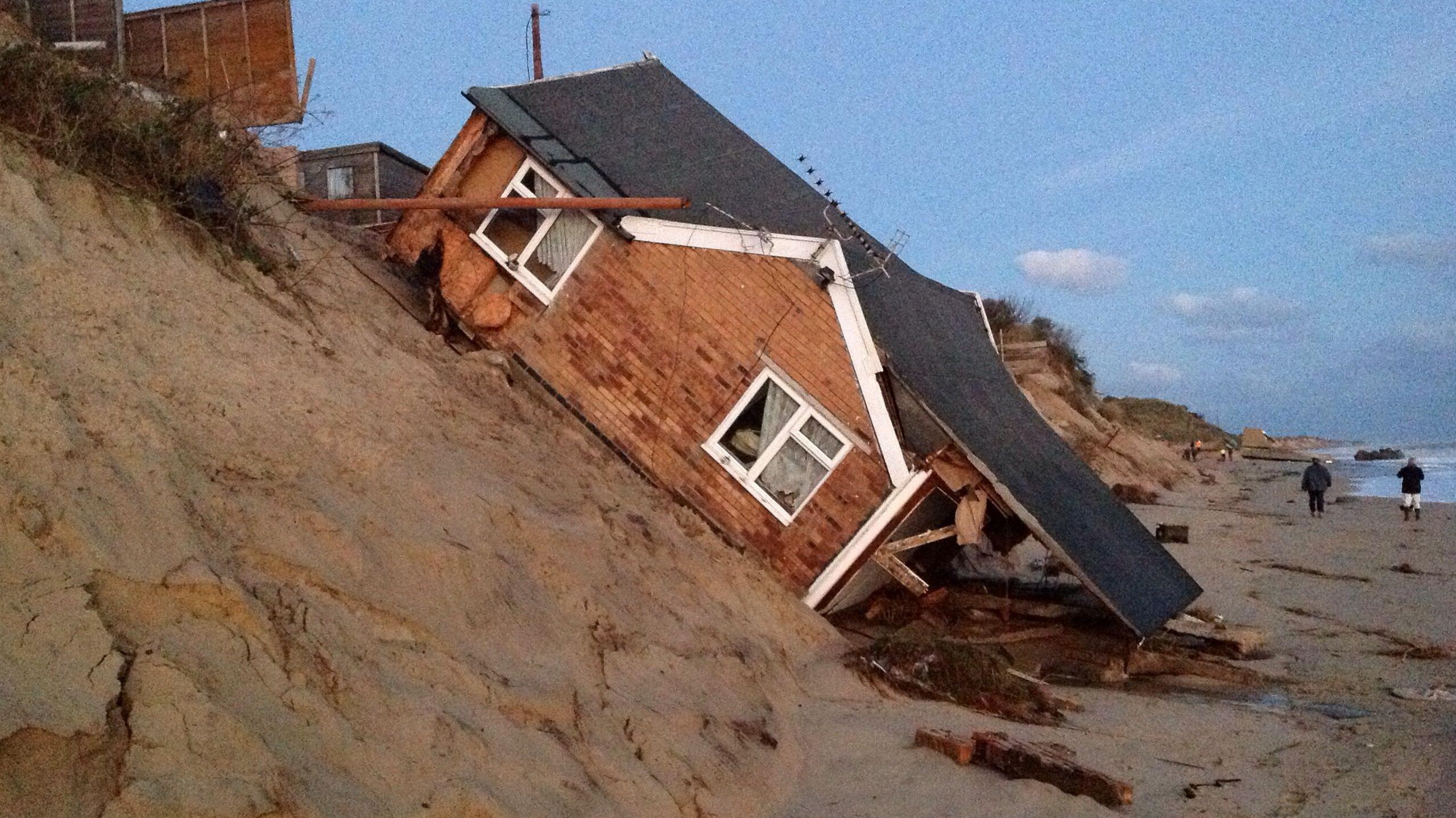 A house is shown in a ruined state, having fallen from a cliff in Hemsby. The roof is partially off and the house itself is resting at a 45 degree angle on sand