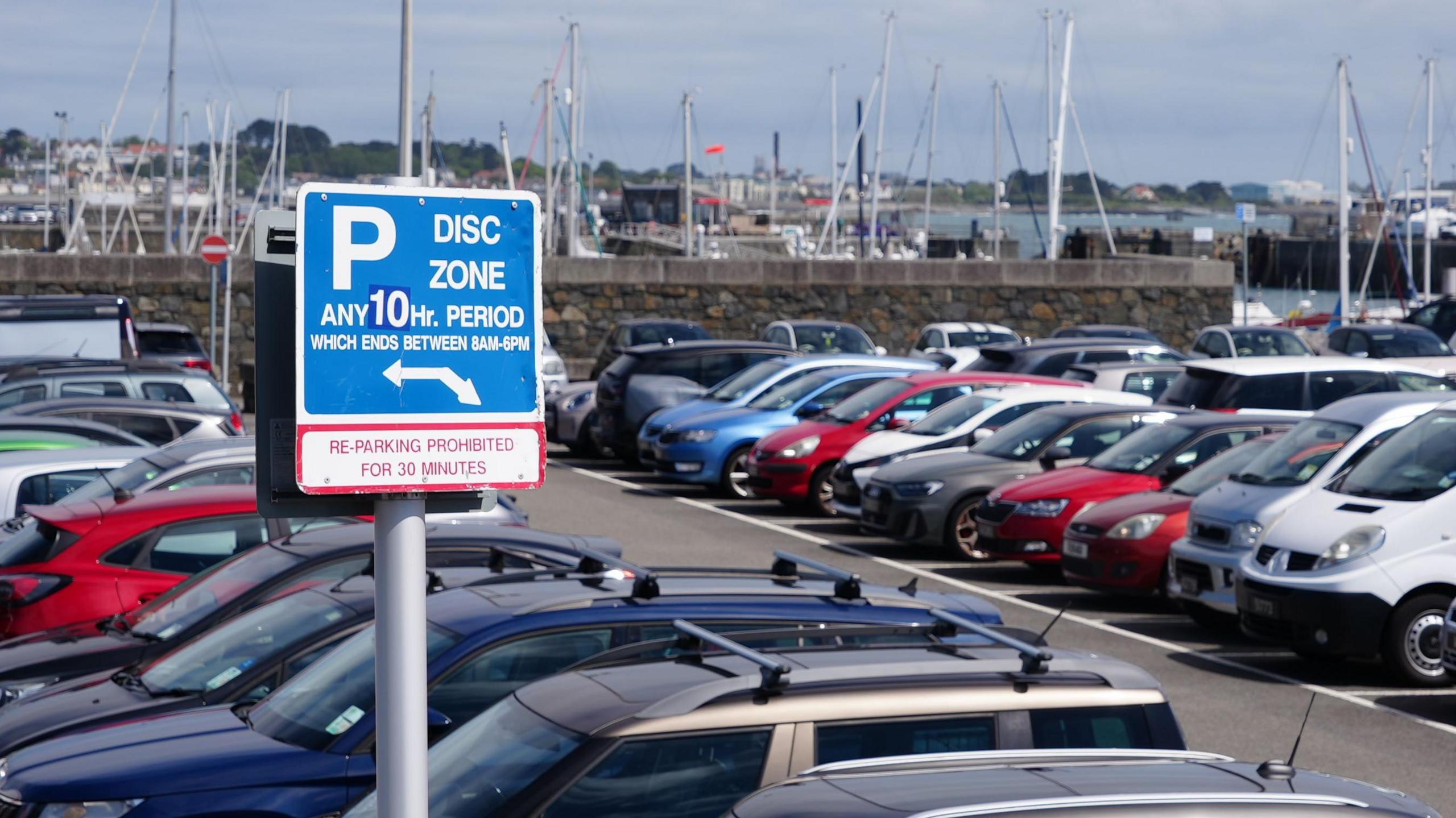 A car park in Guernsey full of vehicles. It is next to a harbour which has several boats docked. A blue sign saying the car park is a disc zone area for any 10-hopur period which ends between 8am and 6pm is in the car park.