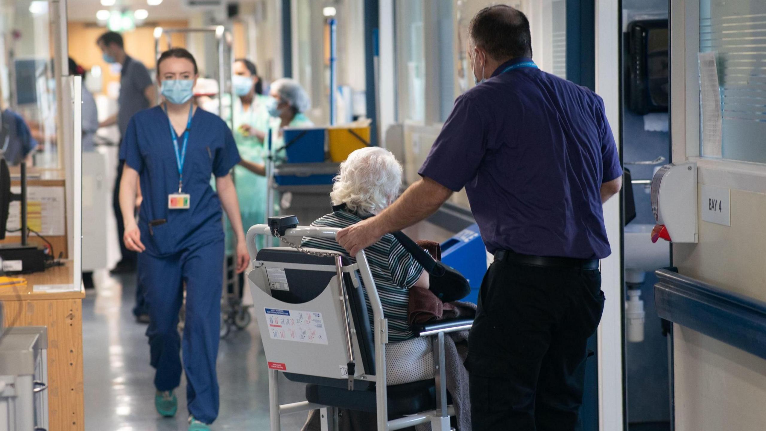 An elderly woman sits in a wheelchair in a hospital ward. A staff member in blue scrubs walks towards her, while a man in a polo shirt holds the chair.