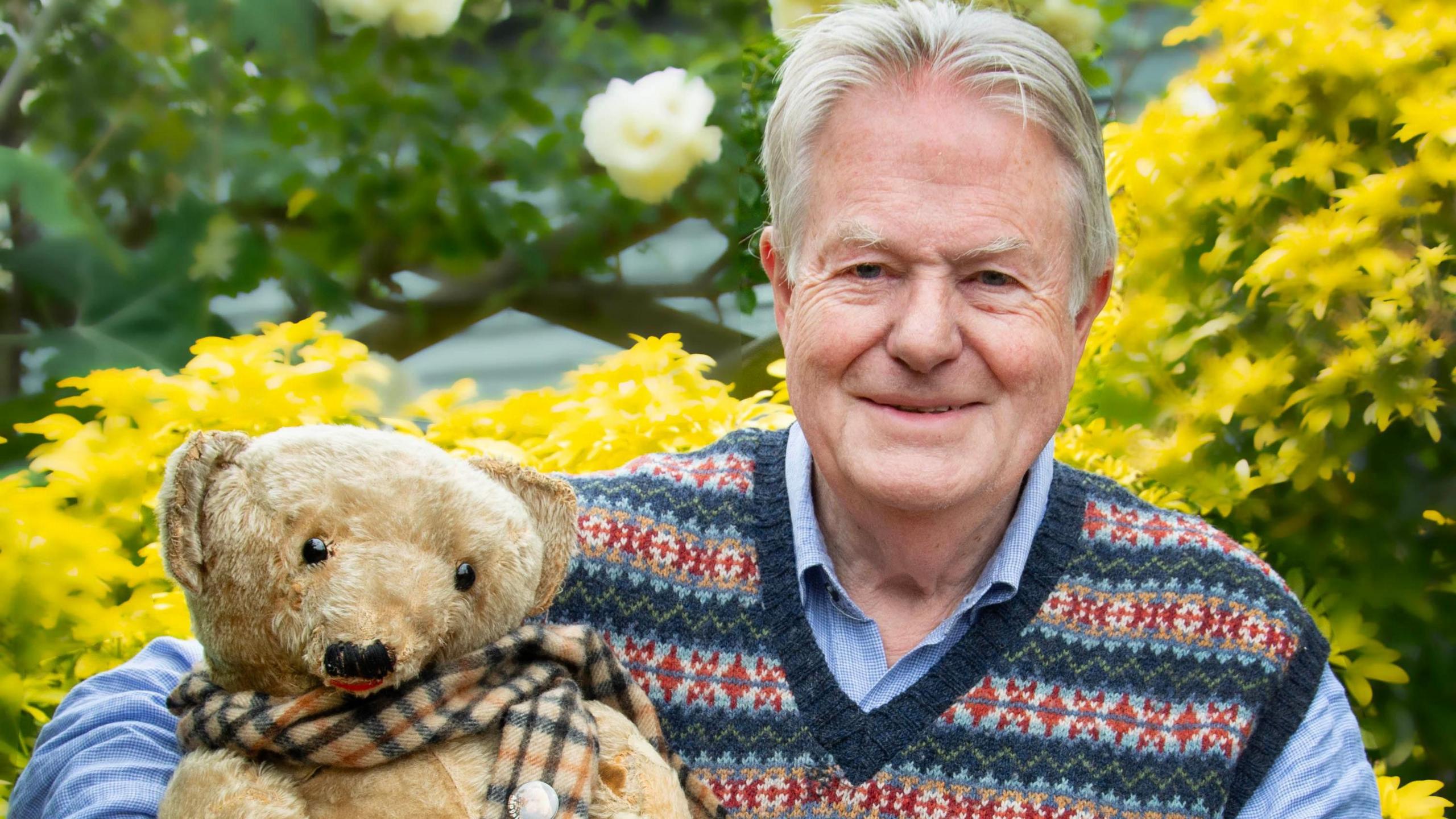 Ian Pout has short combed back grey hear. He is wearing a colourful v-neck jumper over a blue shirt. He is holding an aged-looking teddy bear with a Burberry-style tartan scarf around its neck.