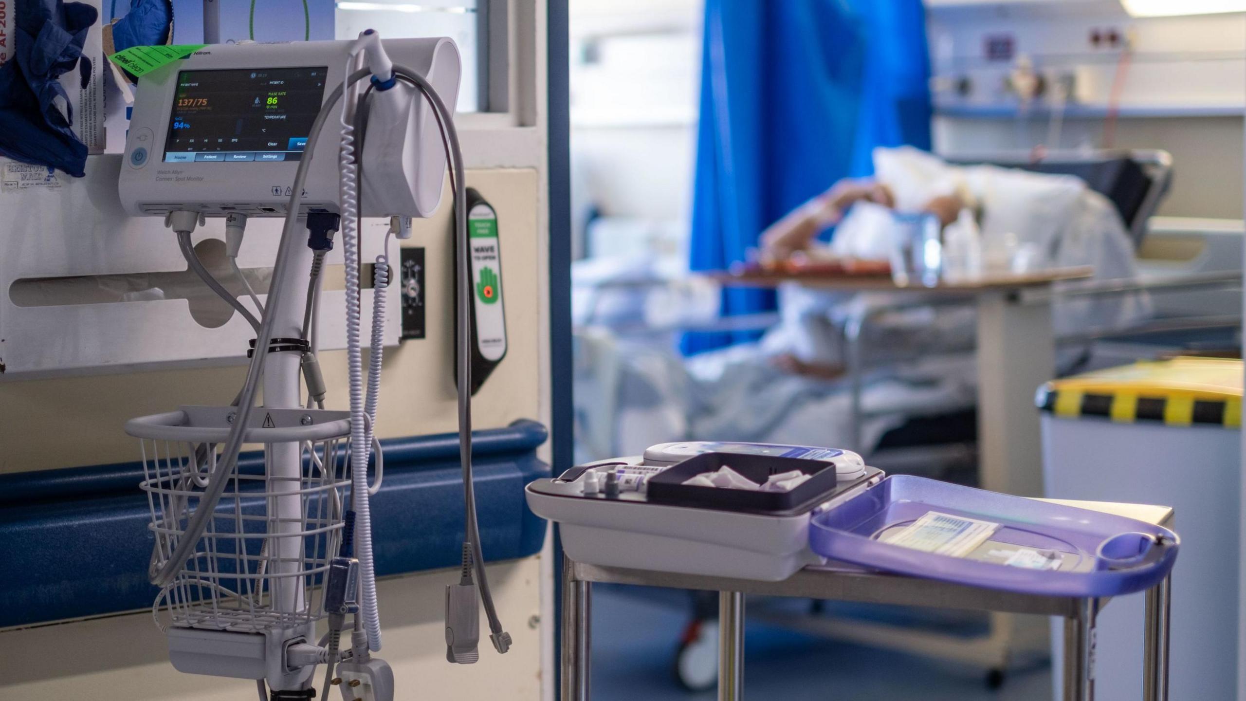 Hospital equipment in a hallway near the entrance of a ward. A patient laying down on a bed with their arm going towards their head can be seen in the blurred background.