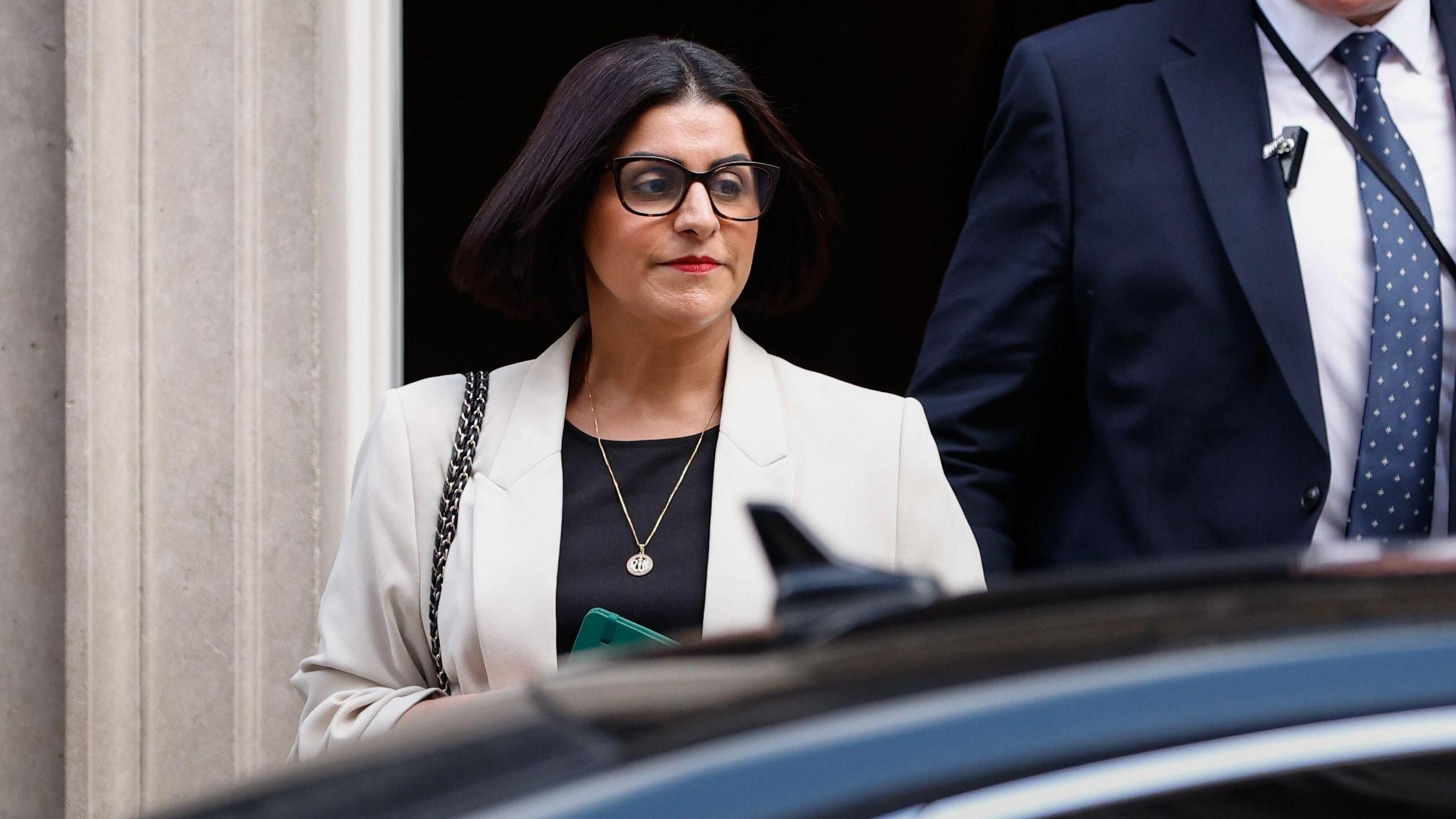 Home Secretary Shabana Mahmood walking to a car wearing dark brown glasses, red lipstick, and a light grey blazer. 