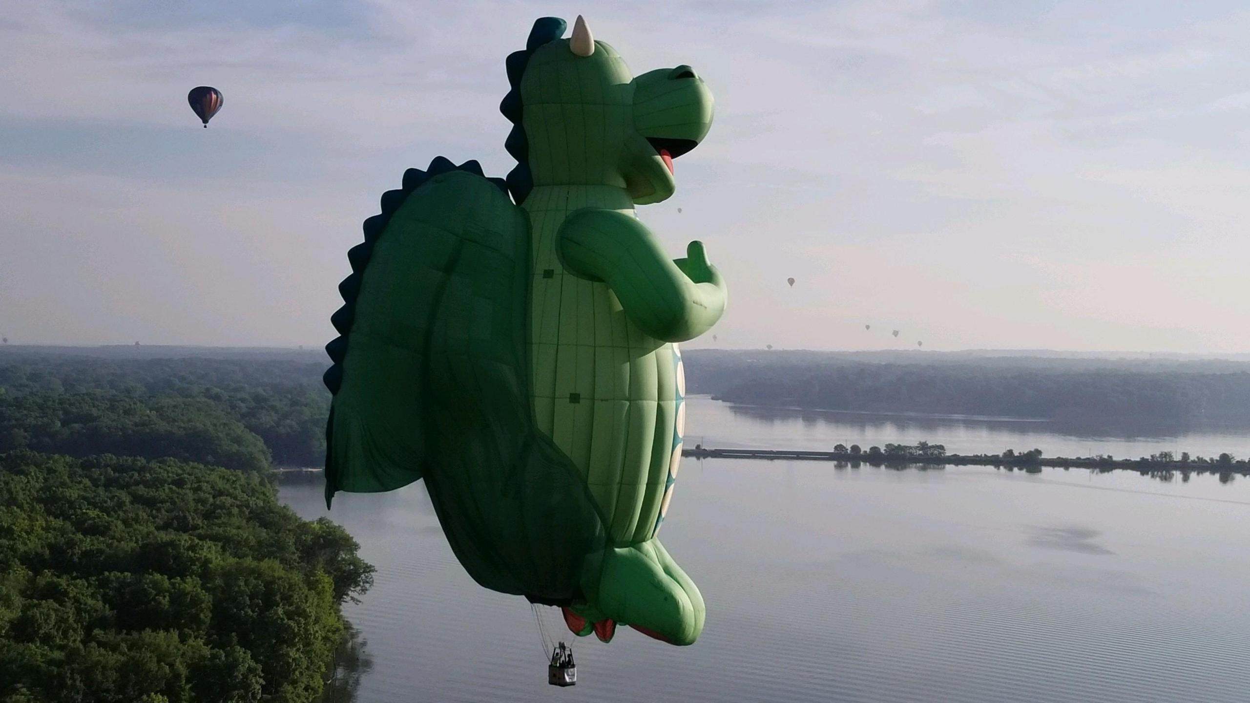 A huge balloon in the shape of a green dragon flies over a river in a hazy day, with smaller balloons flying in the background