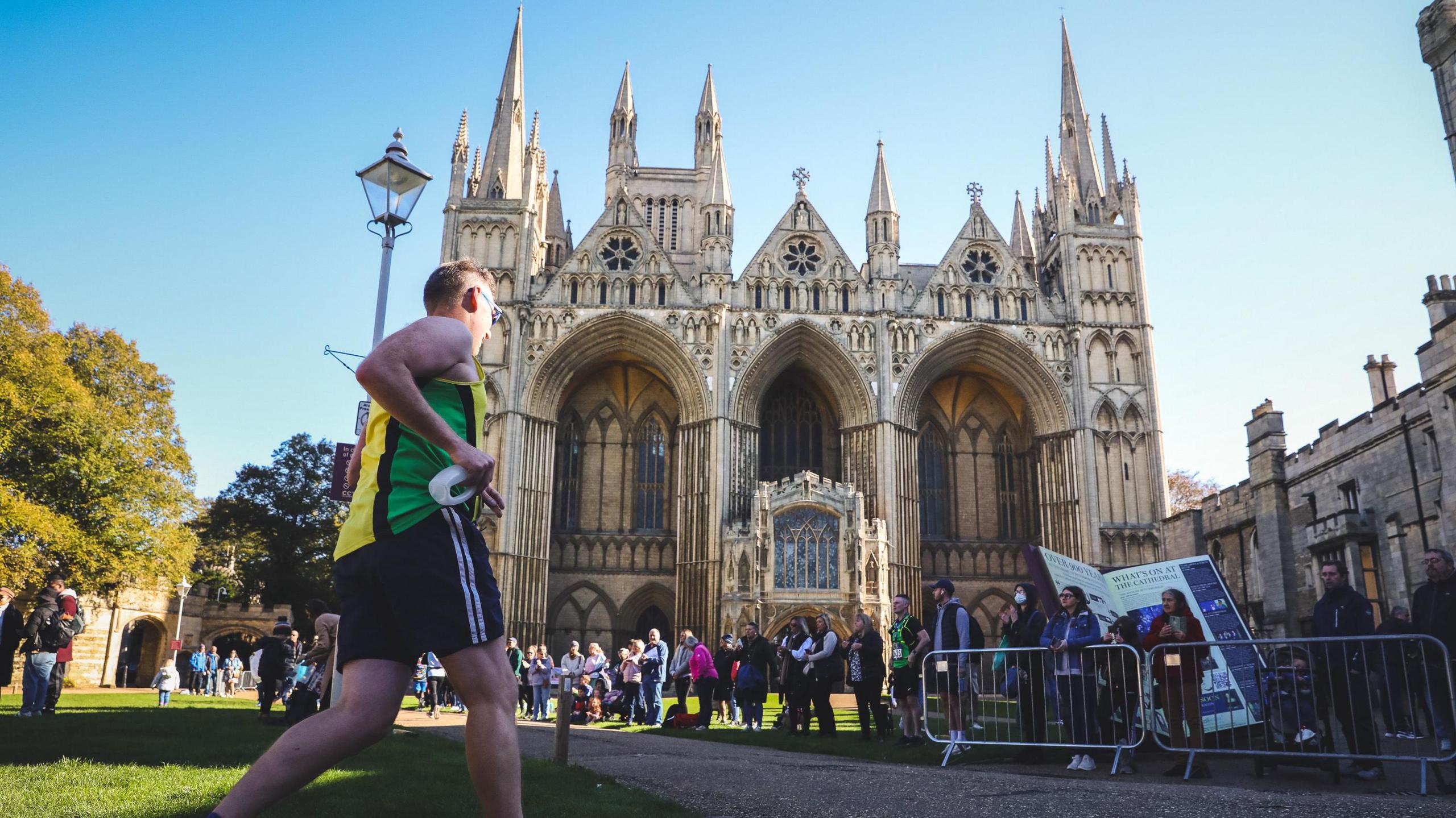 A runner in a yellow and green vest and black shorts running towards Peterborough Cathedral with people lining the path.