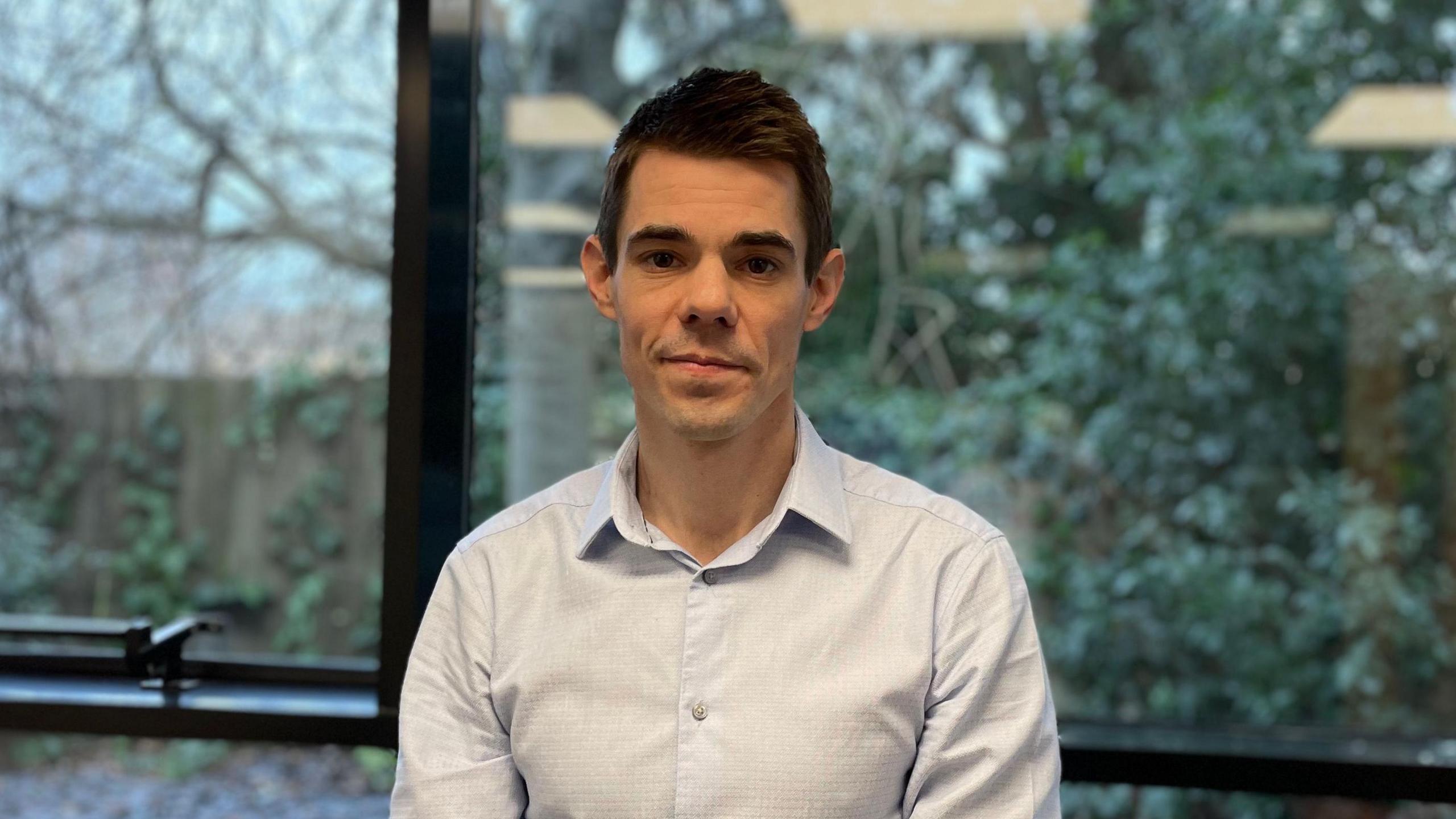 Dan Sexton is standing in an office room, wearing a light blue shirt and standing behind a window, in the background there are trees and bushes.