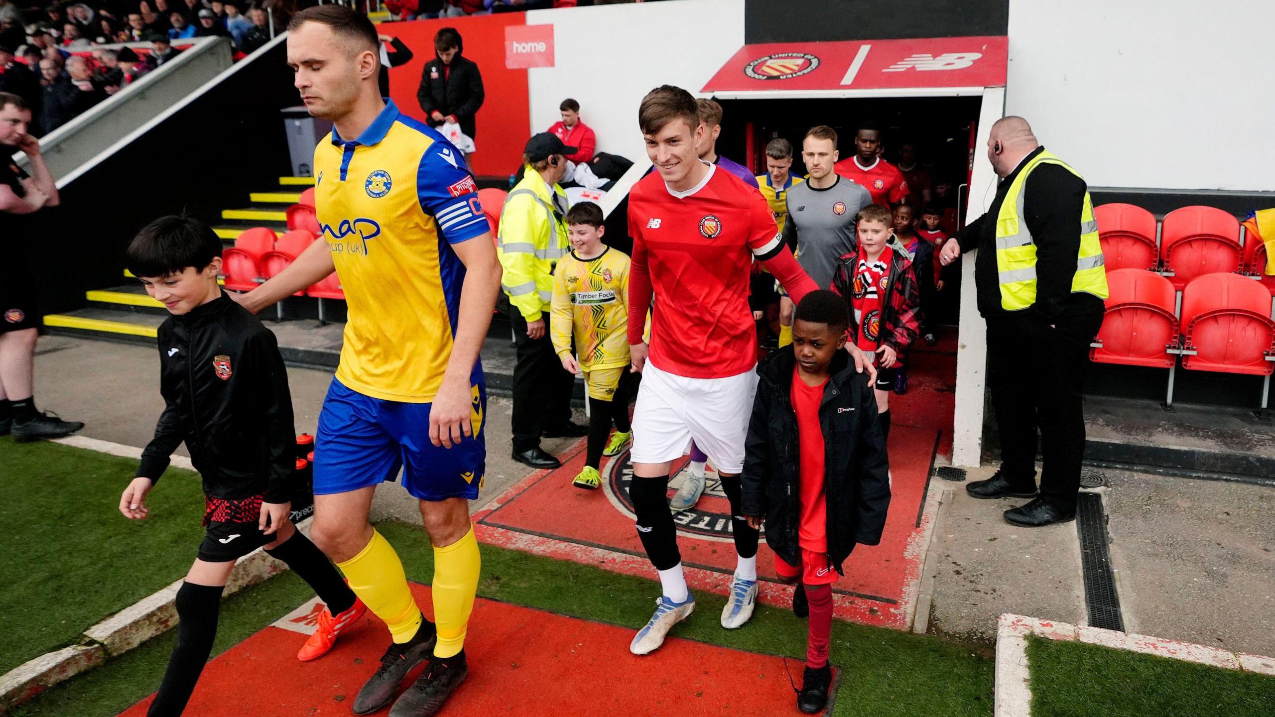 Players from Stockton Town FC and FC United of Manchester walk on to the pitch from the tunnel. Stockton Captain FC Adam Nicholson is leading his team, and is wearing the club's yellow and blue strip. He has arm on the shoulder of a ball boy walking besides him.