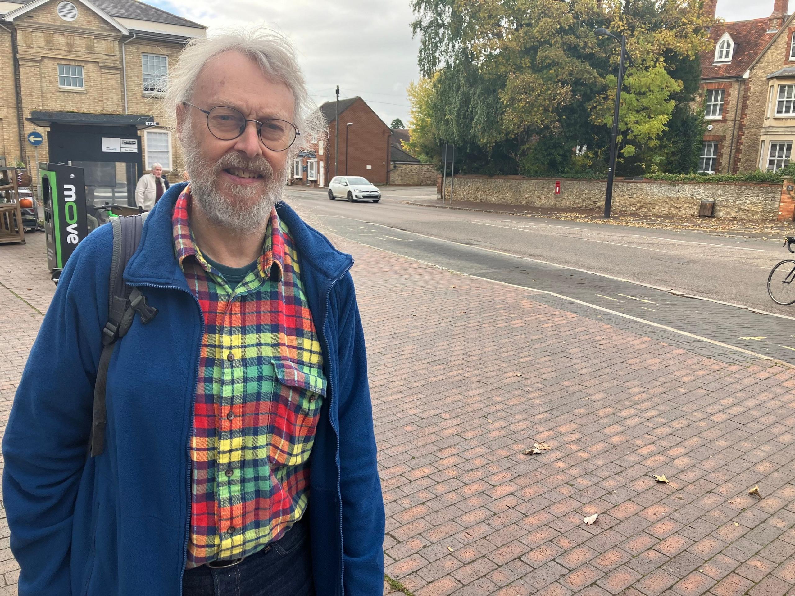 Alan Francis, a middle aged man, with his grey hair tied back in a ponytail, standing on a pavement with the road behind him. He is wearing a blue fleece jacket and a red, yellow and green check shirt underneath. He is stood near a bus stop in Newport Pagnell.