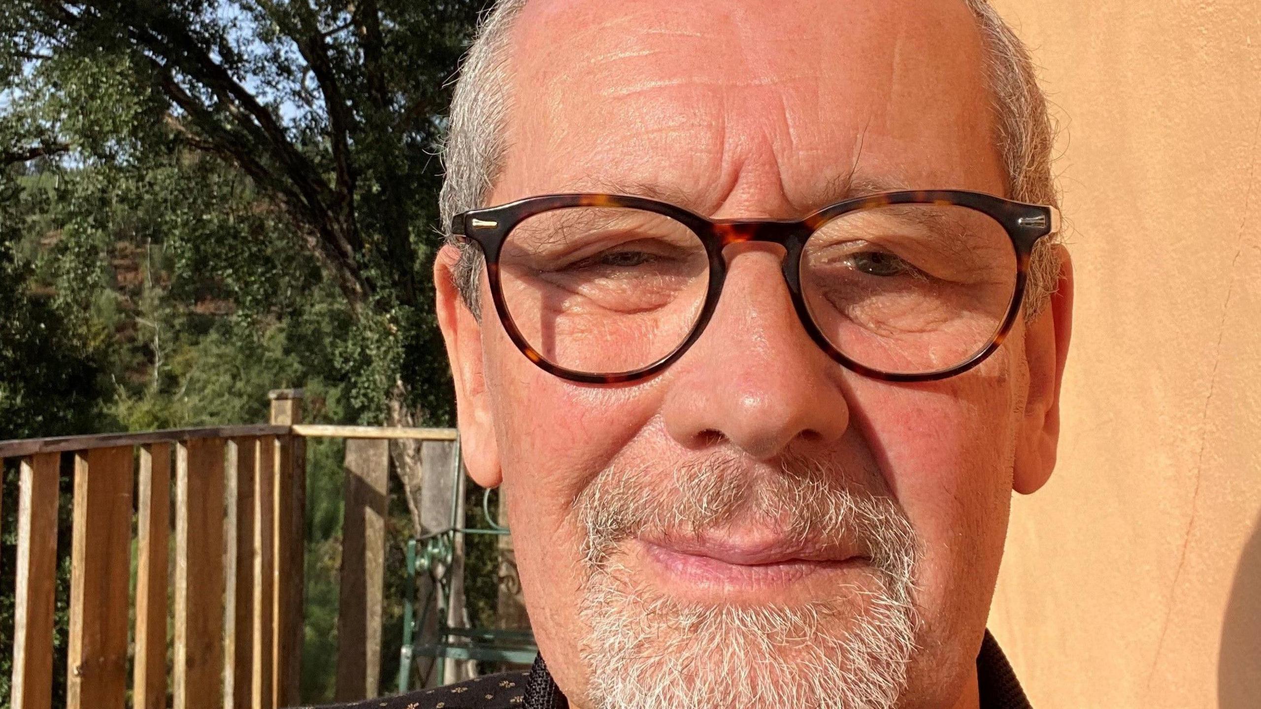 Steve Rice with dark rimmed glasses and a goatee, smiling at the camera outside on a porch