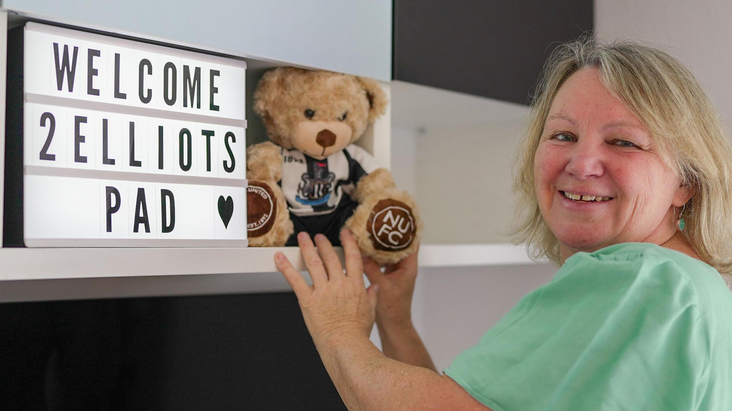 A woman with blonde shoulder-length hair looks over her shoulder and smiles at the camera. She is wearing a pale green tee shirt and holding a small teddy bear wearing Newcastle United kit on a shelf. Next to the bear is an illuminated sign which says 'WELCOME 2 ELIOTS PAD' in capital letters