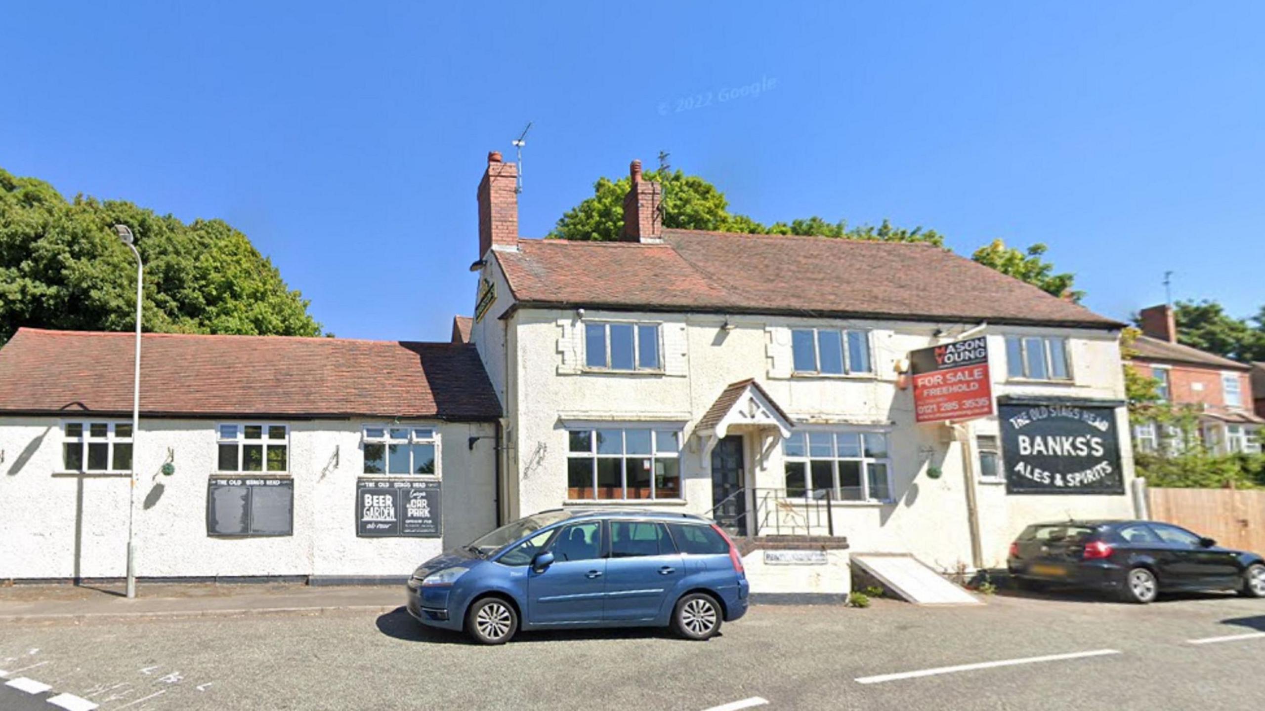 The exterior of a pub with white walls and a black sign on it saying 'The Old Stag's Head, Banks's Ales & Spirits'. A red for sale sign is attached to the building. Two cars are parked outside the building.