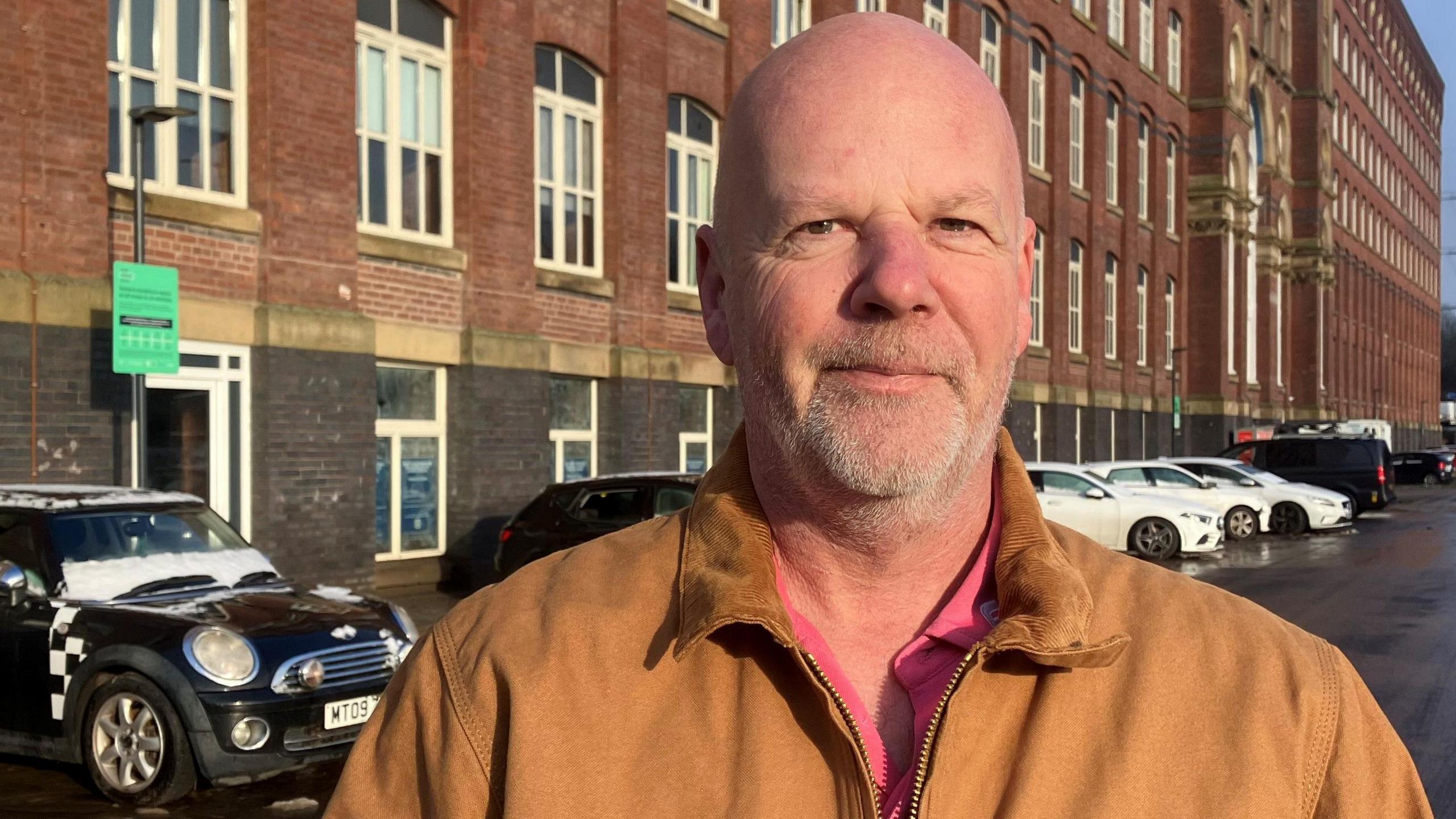 Dave Howarth wearing a tan jacket and salmon-coloured shirt stands in the car park outside the entrance to the former cotton mill, Meadow Mill, in Stockport, where he lives in a rented apartment. 