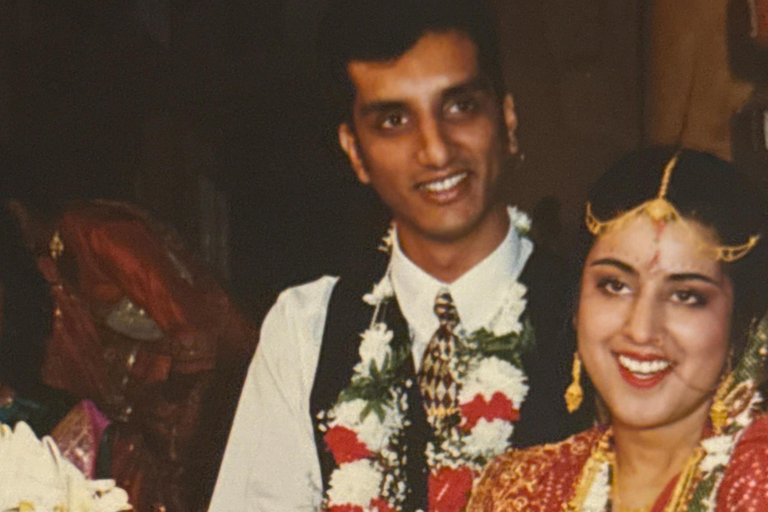 Sarju and his wife are pictured on their wedding day, cutting a three tiered wedding cake. His wife is wearing an intricately designed Panetar, the traditional white and red Gujarati bridal saree, complemented by a Gharchola, a rich red silk saree with bandhni work often draped over the Panetar as part of Gujarati wedding customs. Sarju is dressed in a white shirt with a black waistcoat and tie. It is a head and shoulders shot, with both looking at the camera.