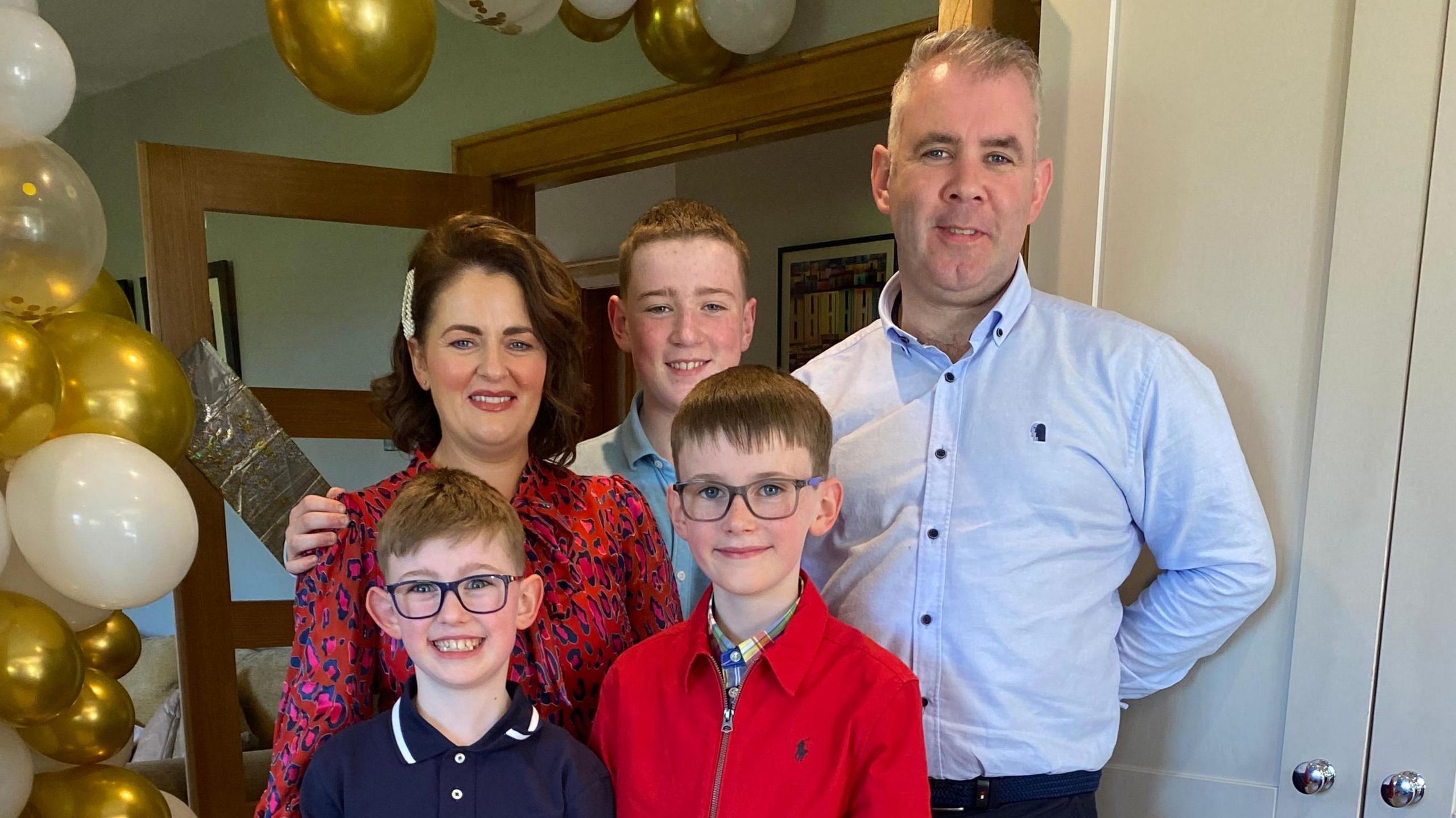 The Sherry Family all stand and pose for a picture in a room with gold and white balloons at the side. Catherine is stood on the left and is wearing a red, blue and pink leopard print dress and has short, shoulder-length brown hair. Beside her is her eldest son who has a blue shirt on and has short light coloured hair. On the right is Fergal, who has short grey hair and is wearing a shirt collared shirt. At the front are the two youngest sons, who both have medium-length brown hair and are wearing dark framed glasses. All of them are smiling at the camera.