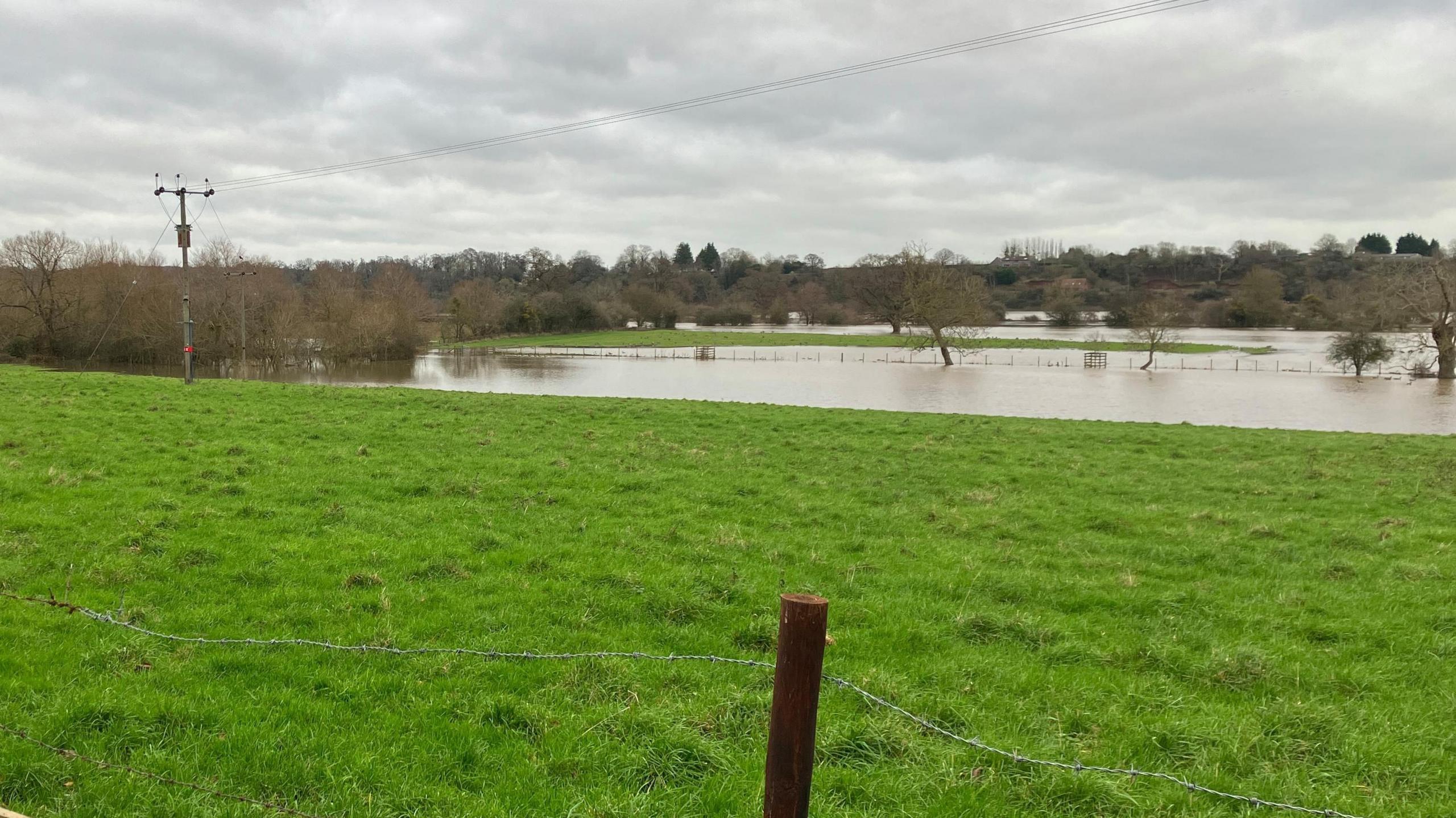 A field which has been partially flooded. Electricity pylons are visible - as they stretch into the distance, some of them can be seen partially submerged.