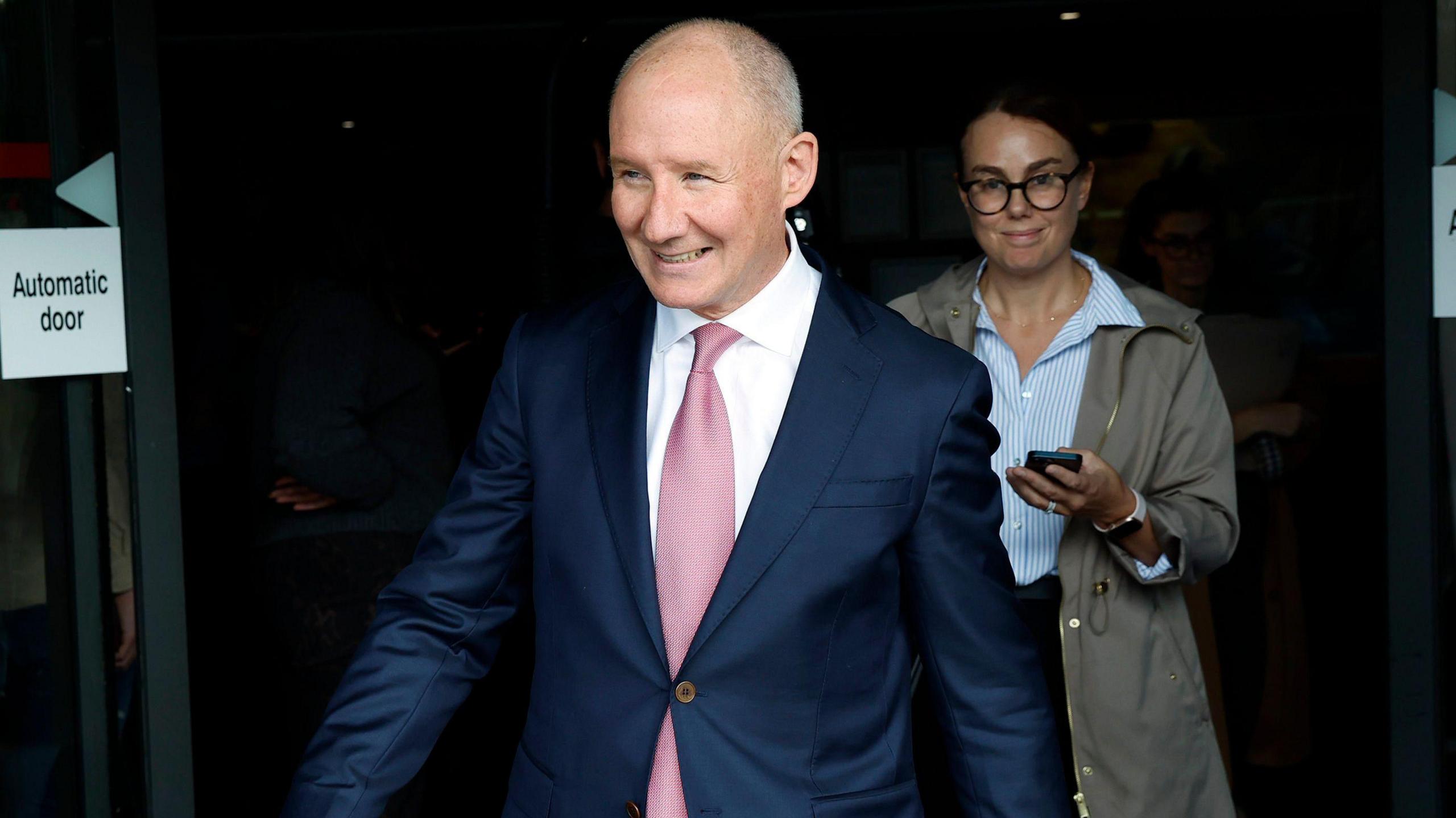 Jim Gavin walks outside of RTÉ in Dublin smiling. He is an older, balding man with short grey hair. He is wearing a dark navy blue suit jacket, a pink tie and a white collared shirt.