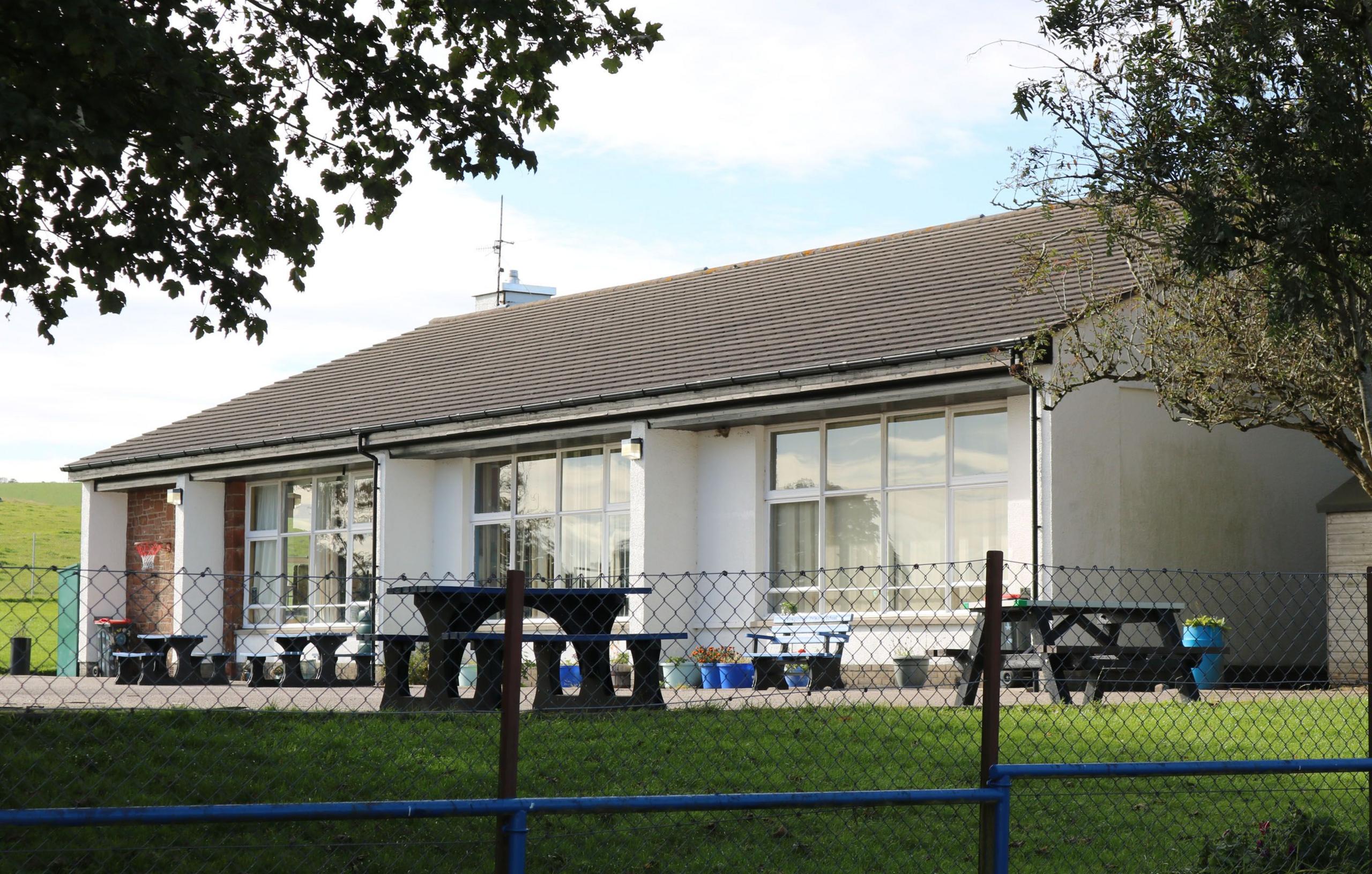 A small school building with a low tiled roof and a white frontage surrounded by picnic tables, grass and a low fence