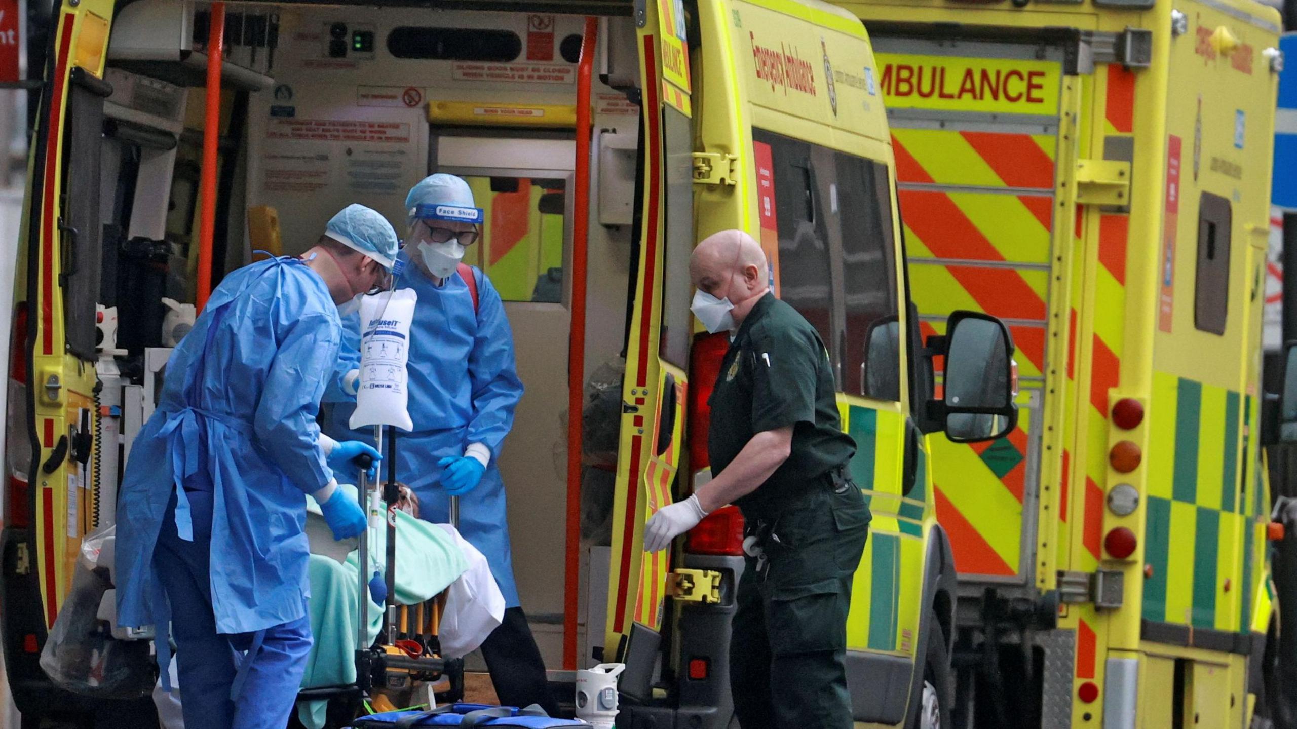 An ambulance with its doors at the back open and two health workers wearing blue clothing and masks and another paramedic wearing green clothing and a mask with a patient on a trolley