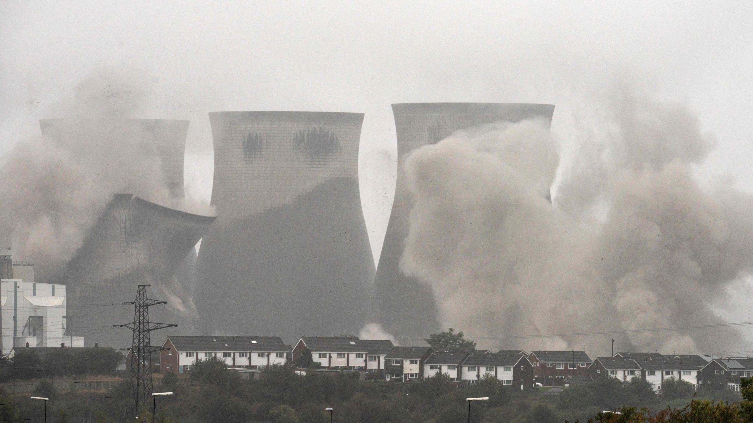 A grainy picture of four large power station cooling towers being demolished in a controlled explosion. One tower is shown partially collapsed amid clouds of smoke. There are several houses in the forefront. 