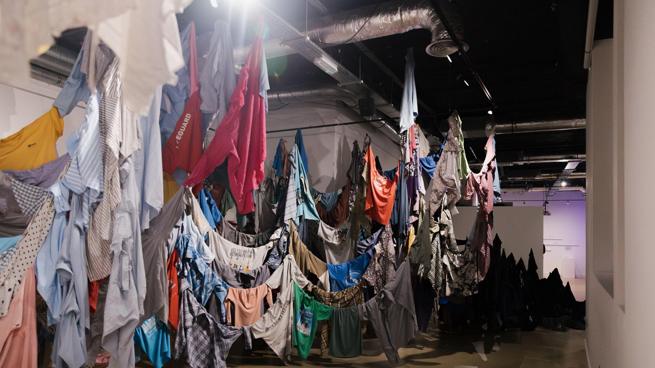 Shirts of different colours, designs and sizes are hung indoors around lines that drop down close to the floor where some of the shirts are touching the floor. There are hundreds of shirts on display.